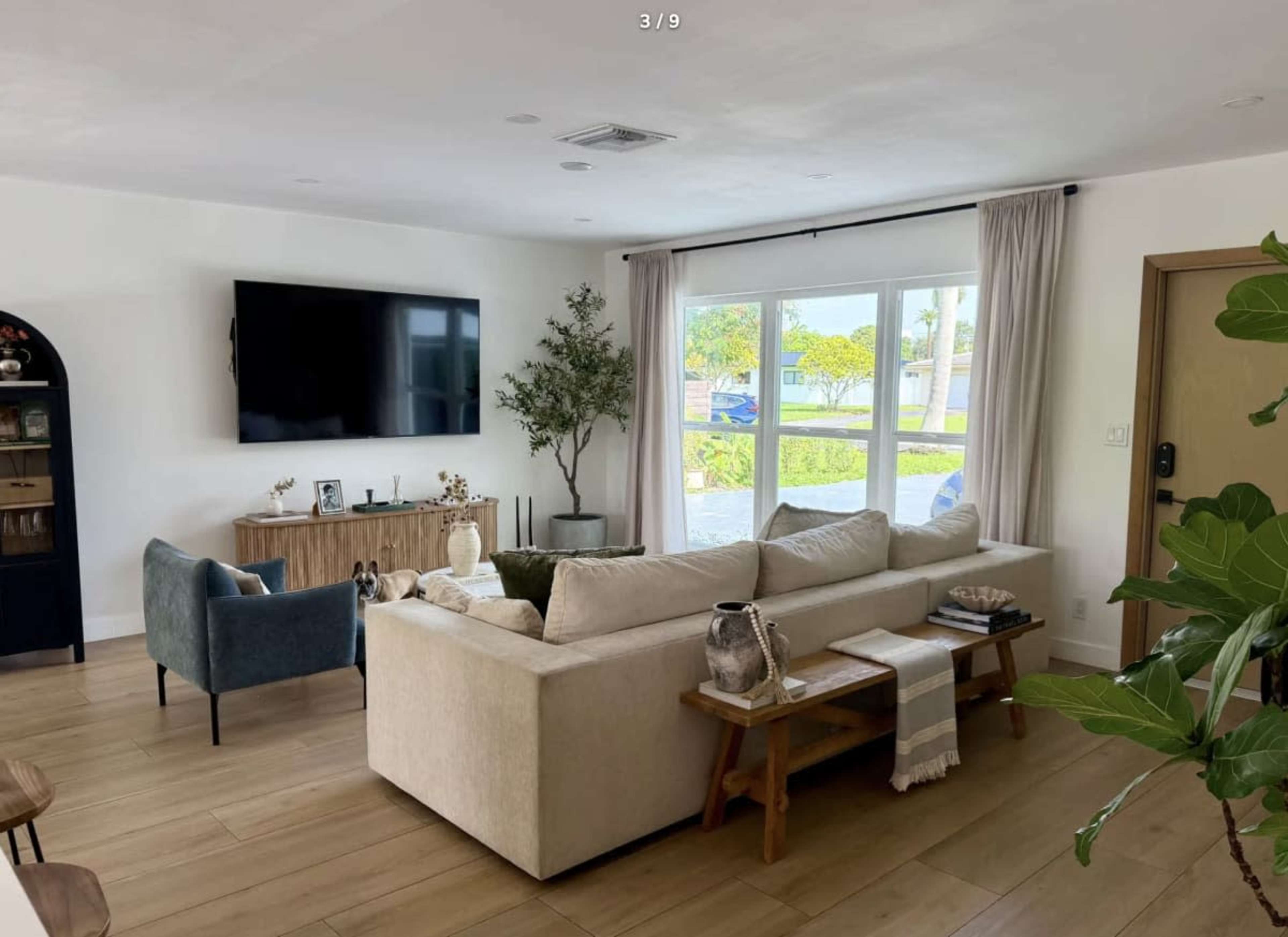 The image shows a modern living room with a light-colored sectional sofa, a wooden coffee table, a flat-screen TV on the wall, and large windows letting in natural light.