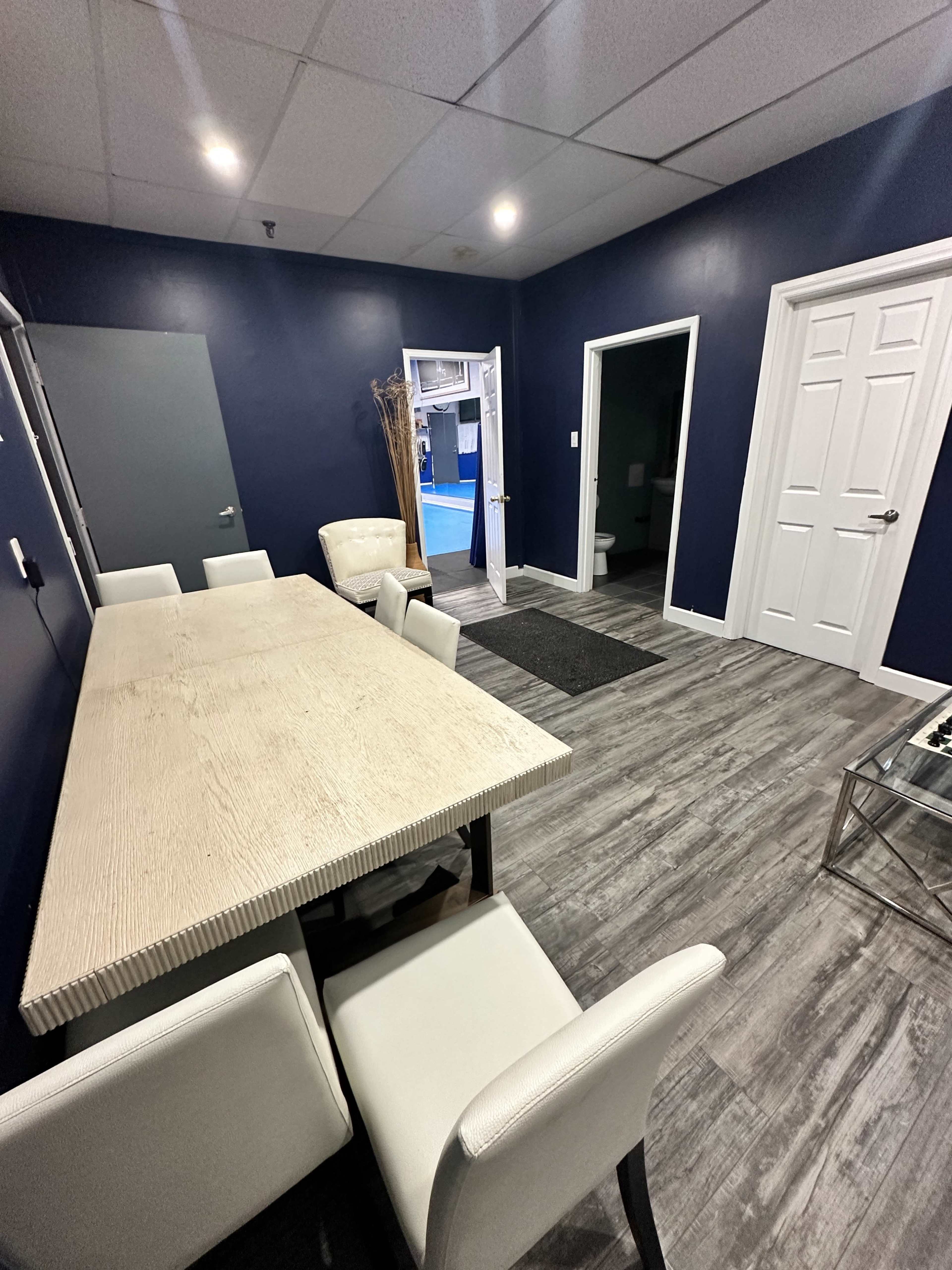 The image shows a modern meeting room with a large table surrounded by white chairs, dark blue walls, and two doors leading to other areas.