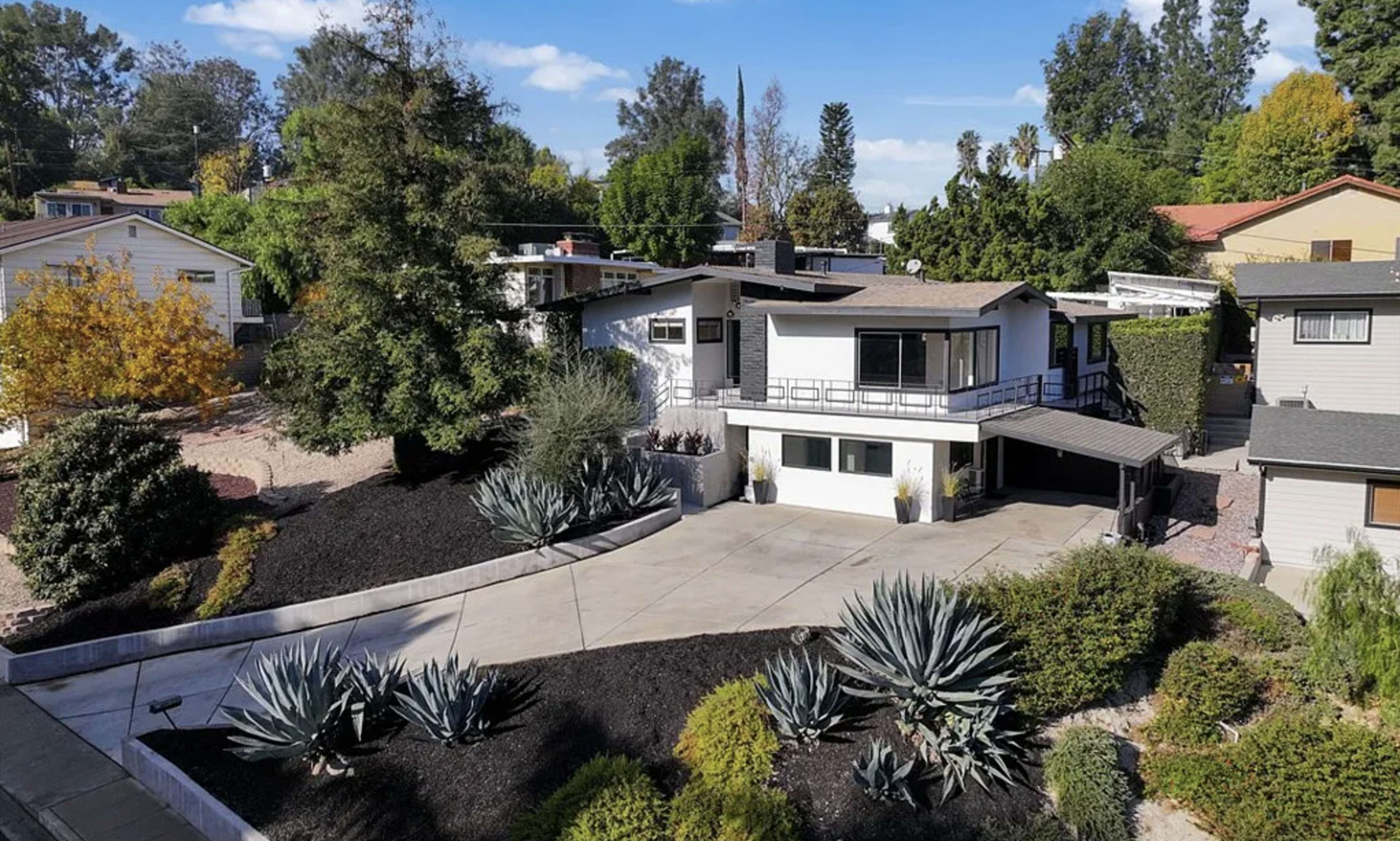 The image shows a modern house surrounded by landscaped gardens and trees, with a paved driveway leading to the entrance.