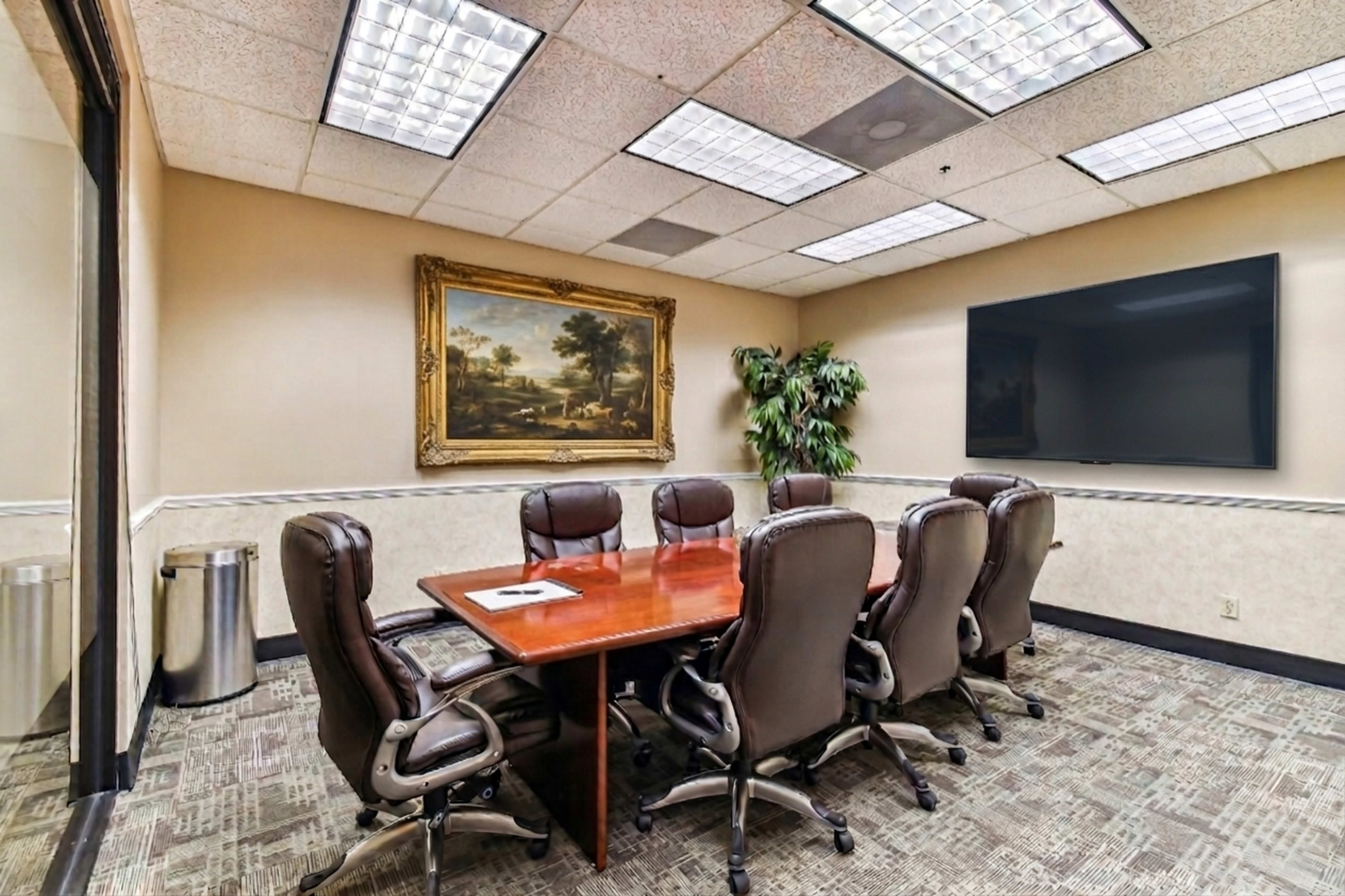 The image shows a conference room with a large wooden table surrounded by eight leather chairs, a framed painting on the wall, and a large television mounted on the opposite wall.
