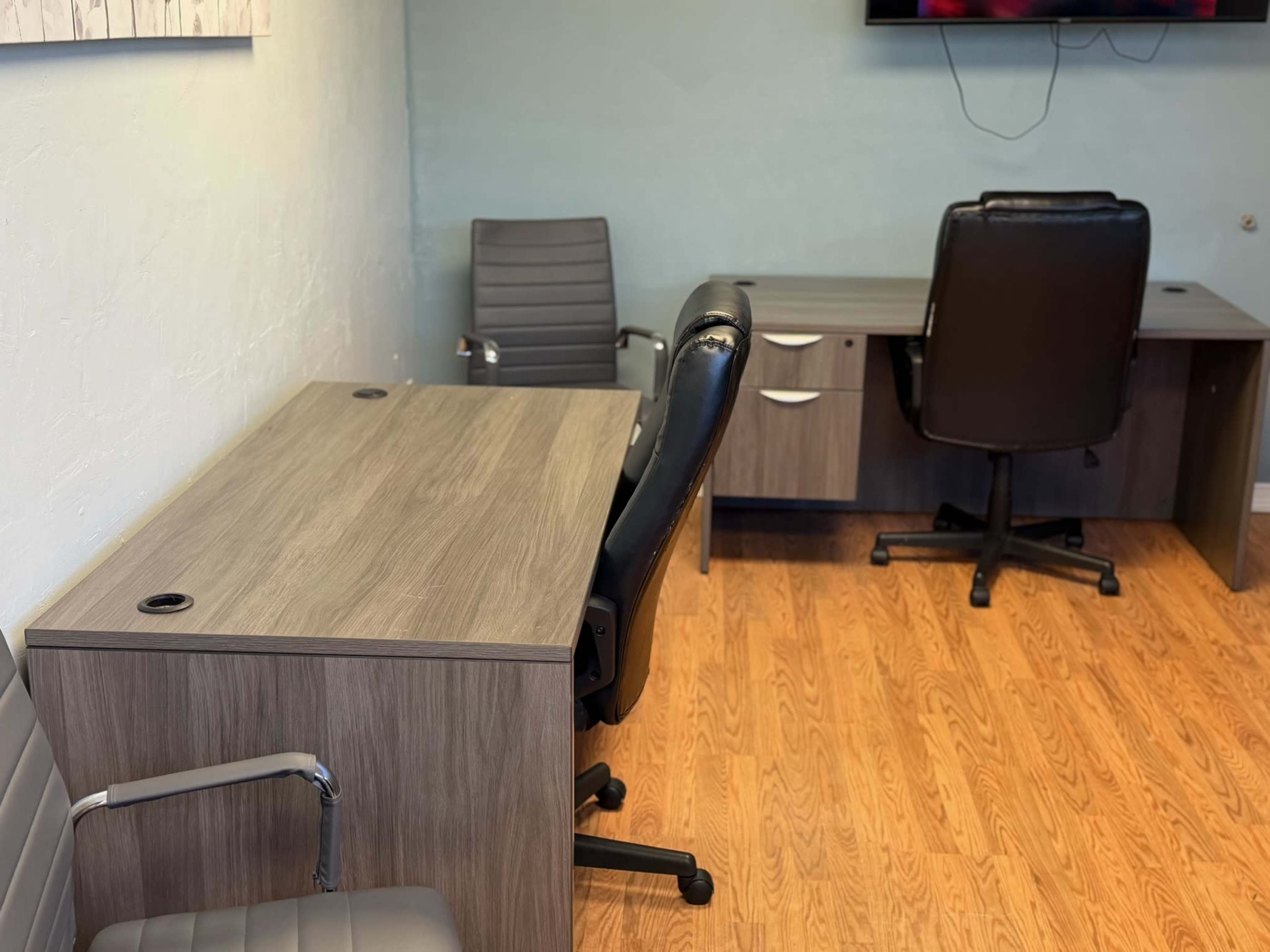 The image shows a small office space with two gray office chairs and a desk made of light wood, accompanied by a television mounted on the wall.