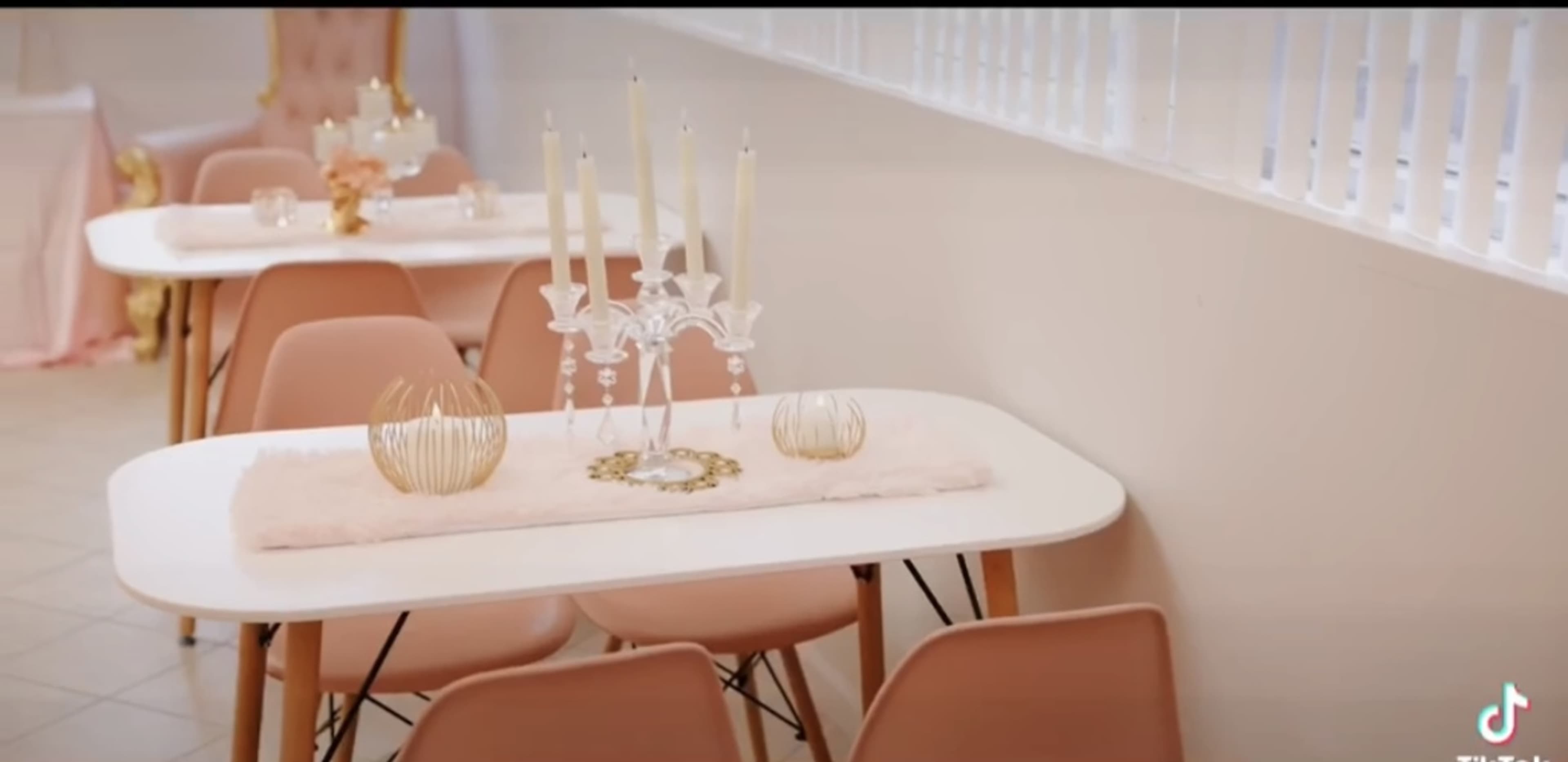 A brightly lit room features elegant tables with soft pink chairs, adorned with decorative candles and gold accents.