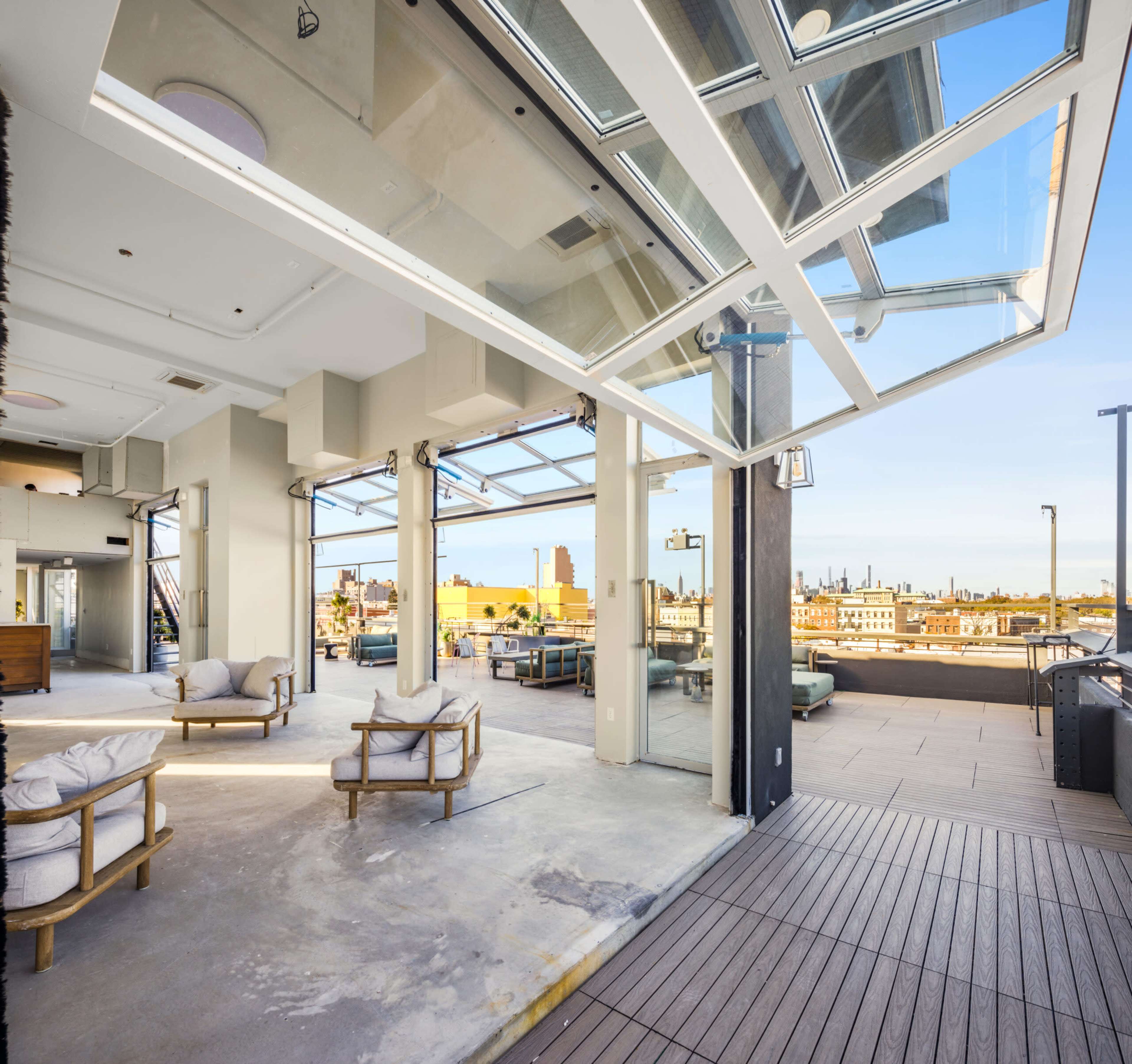 The image shows a spacious, modern terrace with large open windows overlooking a city skyline.