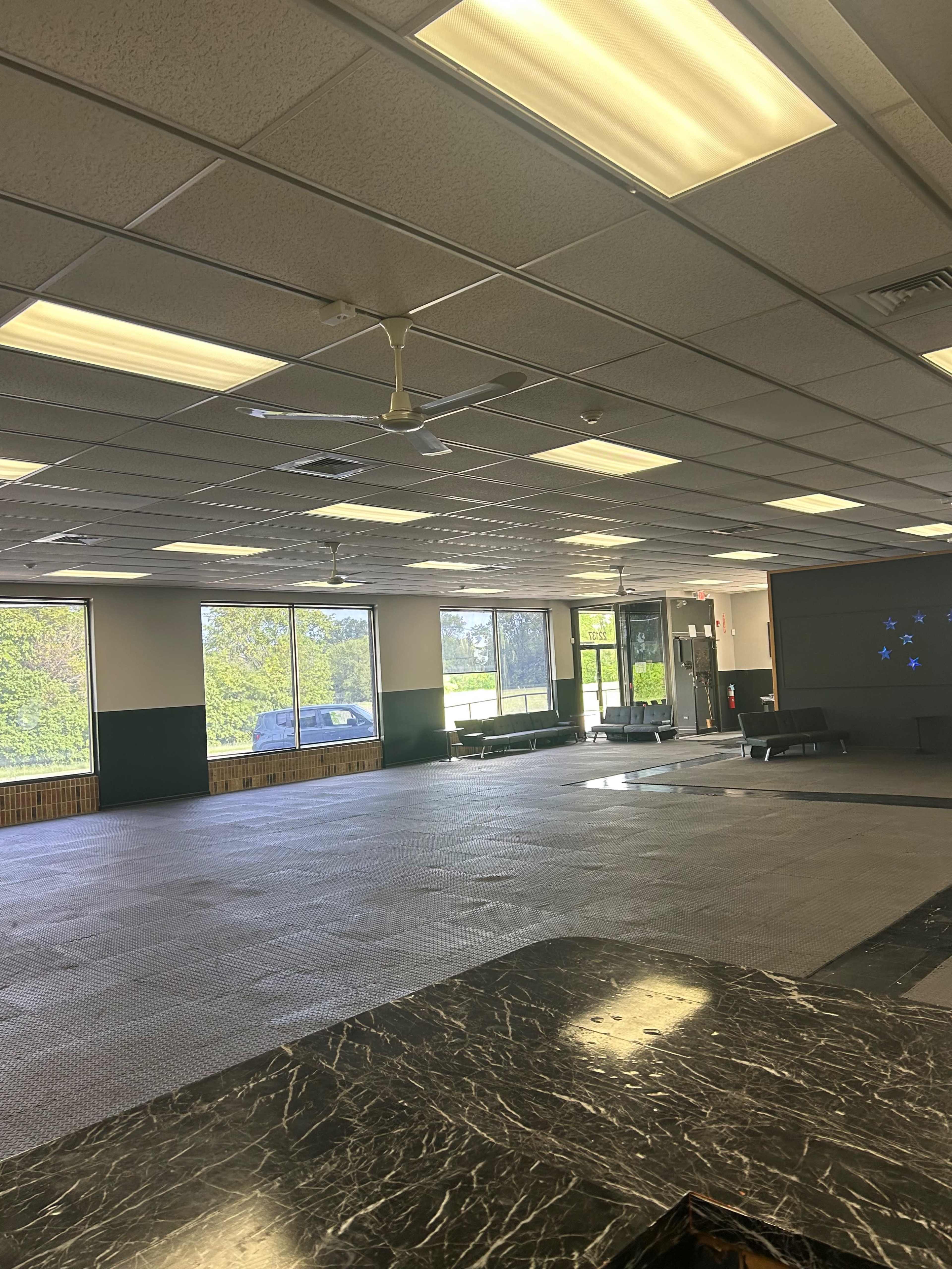 The image shows an empty room with a patterned floor, large windows allowing natural light, and basic overhead lighting.