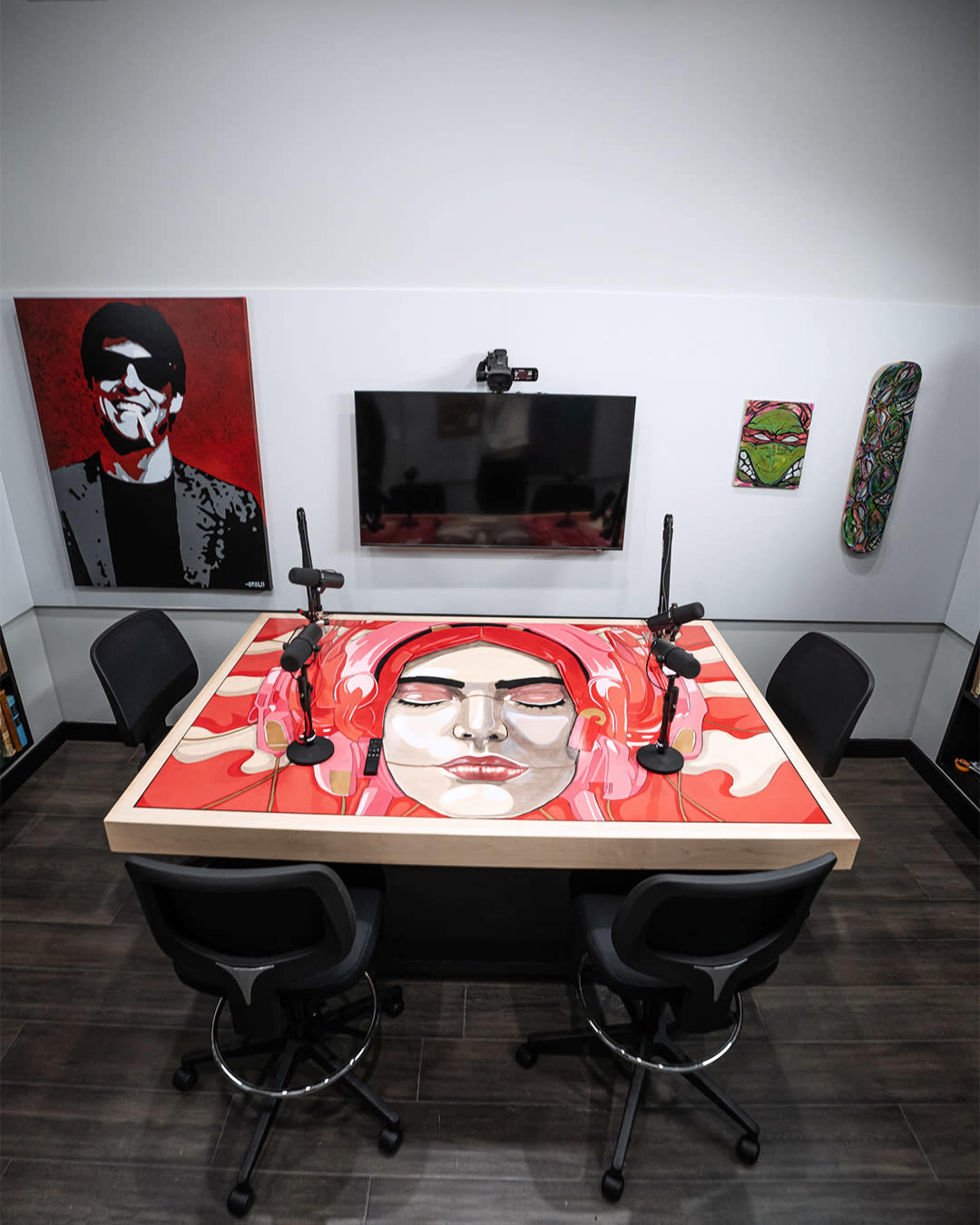 A modern podcast studio with a large table featuring a colorful mural, equipped with microphones and chairs, against a backdrop of wall art.