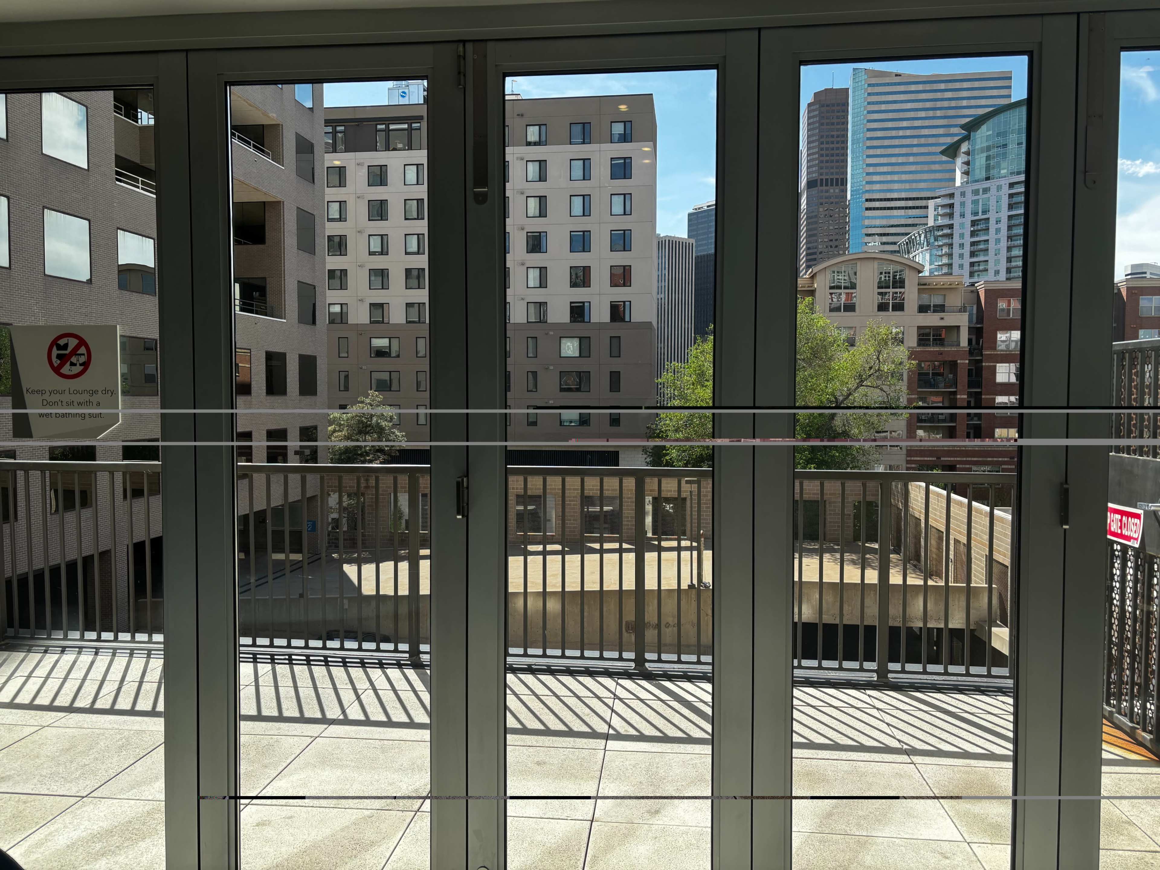The image shows a view from a balcony featuring large glass doors that open to a skyline with modern buildings and greenery outside.