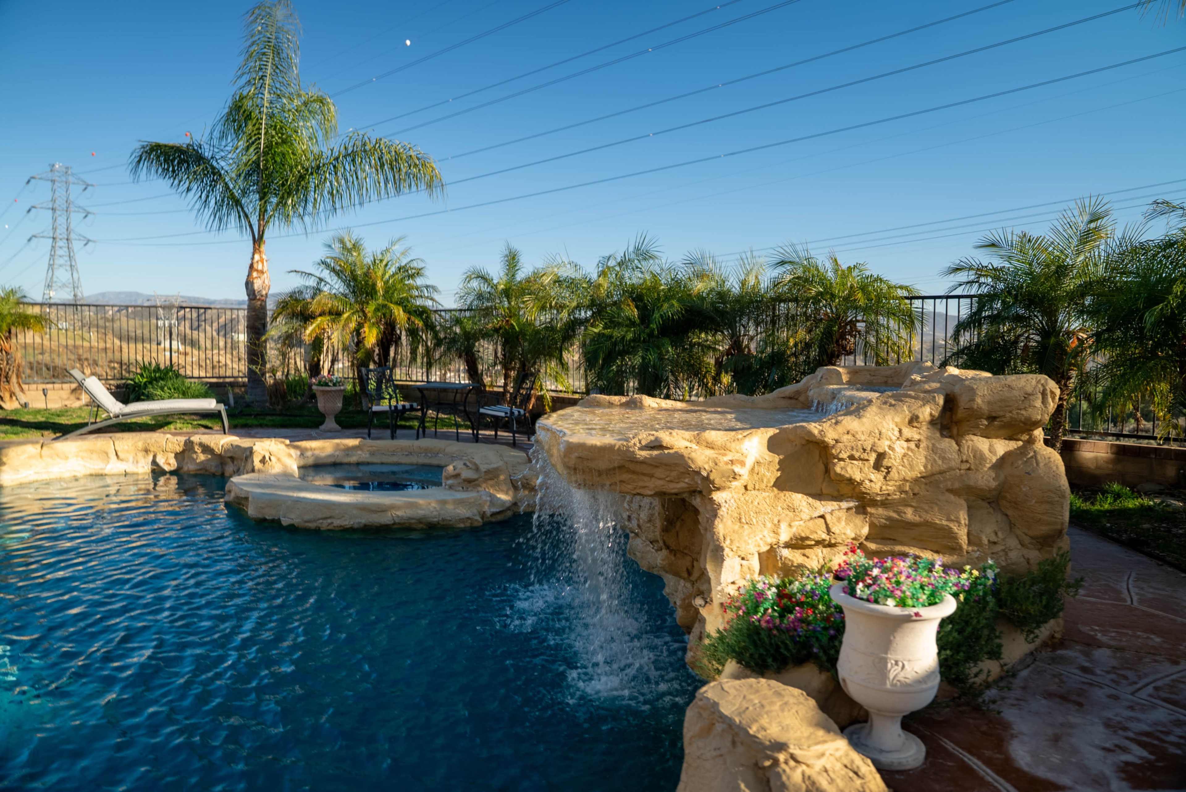 The image depicts a landscaped backyard with a swimming pool featuring a rock waterfall, surrounded by palm trees and outdoor seating.