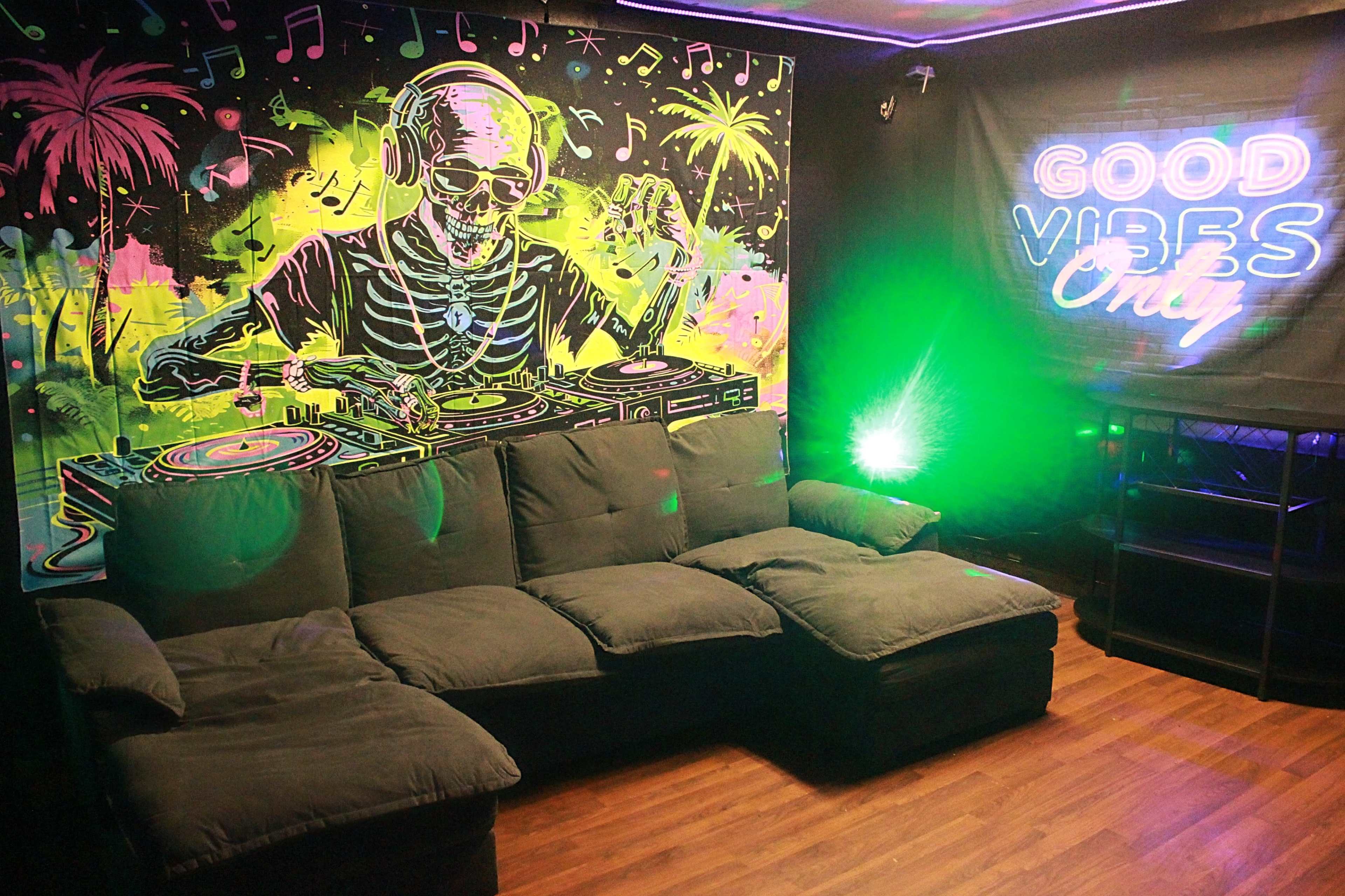 The image shows a vibrant room with a large mural of a DJ skeleton behind a gray sectional couch and a neon sign that says "GOOD VIBES Only."