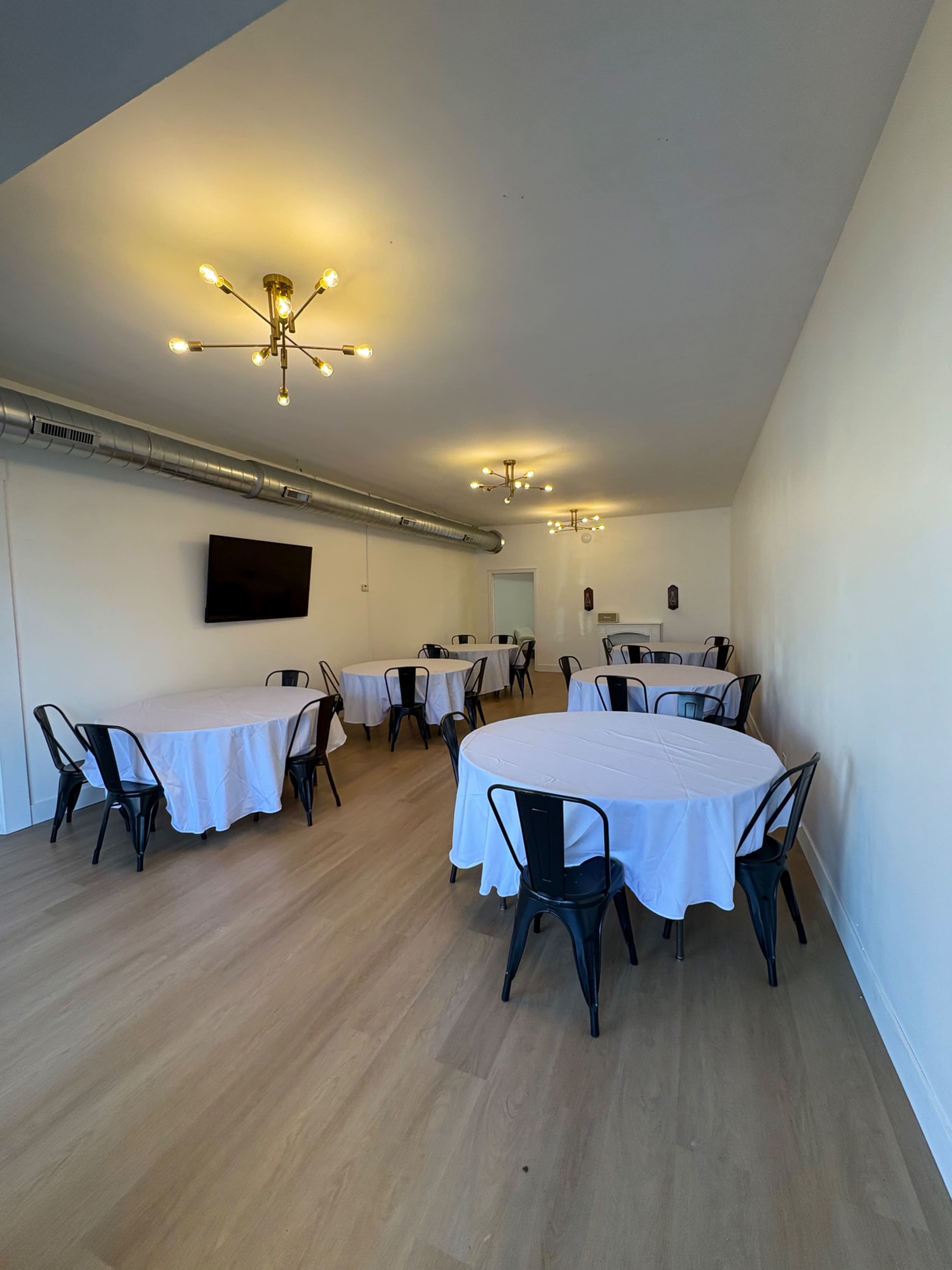 The image shows a well-lit, empty banquet room with several round tables covered in white tablecloths and black metal chairs.