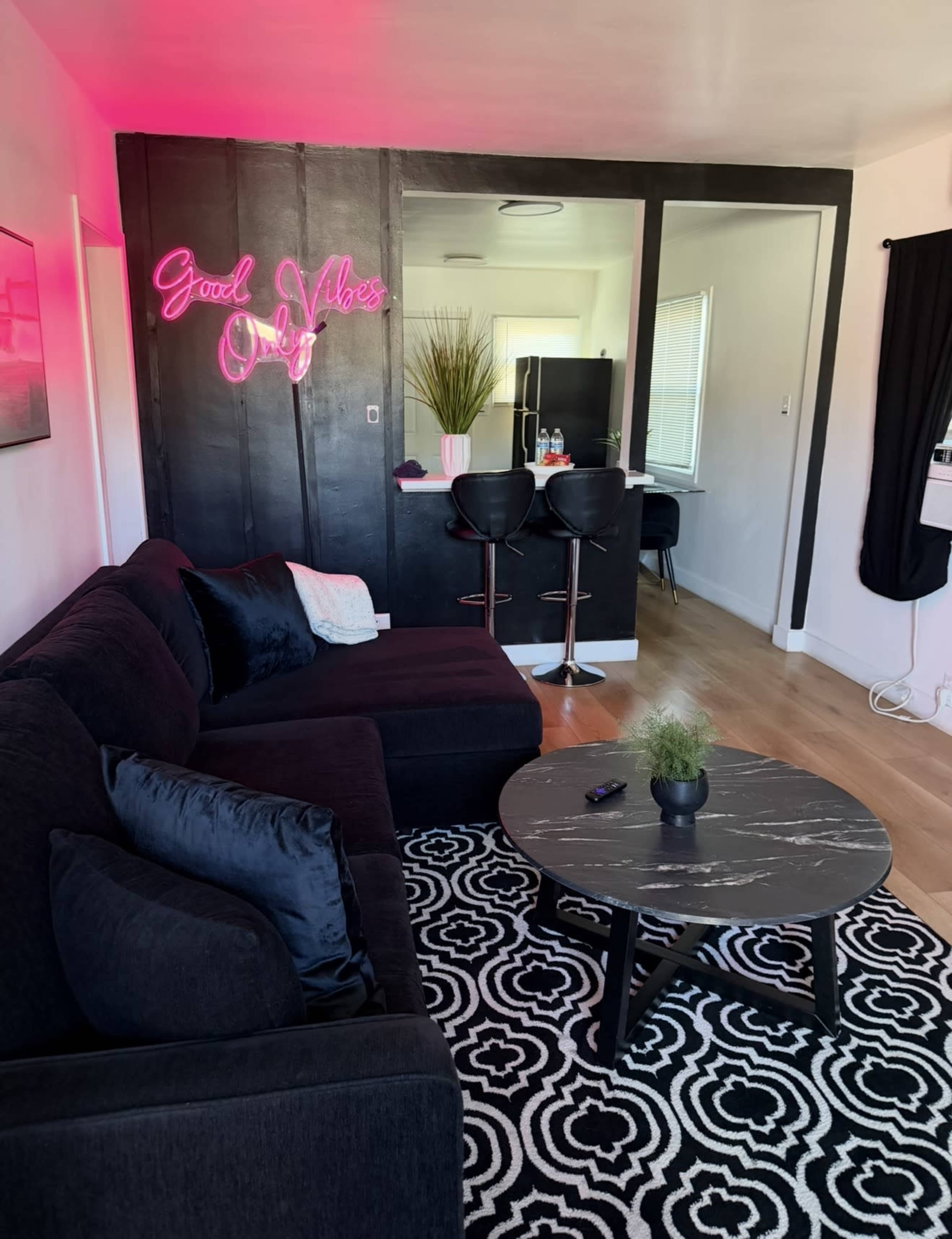 The image shows a modern living room with a black sectional sofa, a round coffee table, and a neon sign that reads "Good Vibes Only" against a black wall, with a glimpse of a kitchenette in the background.