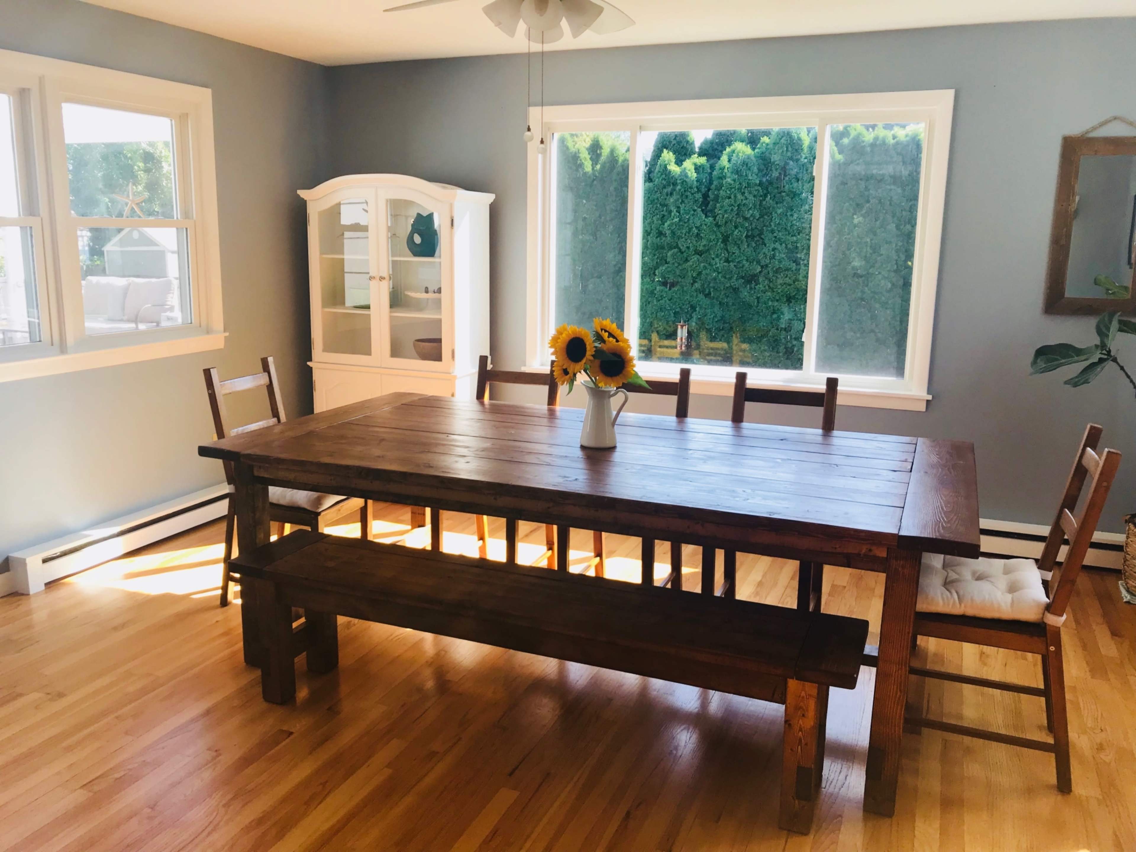 A large wooden dining table with benches and chairs is positioned in a well-lit room featuring a china cabinet and a view of green shrubs through the windows.