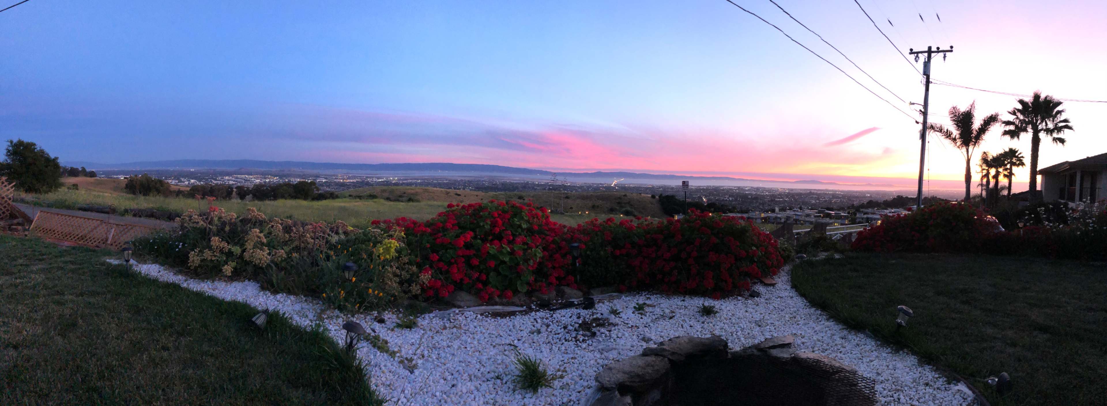 A panoramic view shows a landscaped area with flowers and a pond, overlooking a valley and distant hills at sunset.