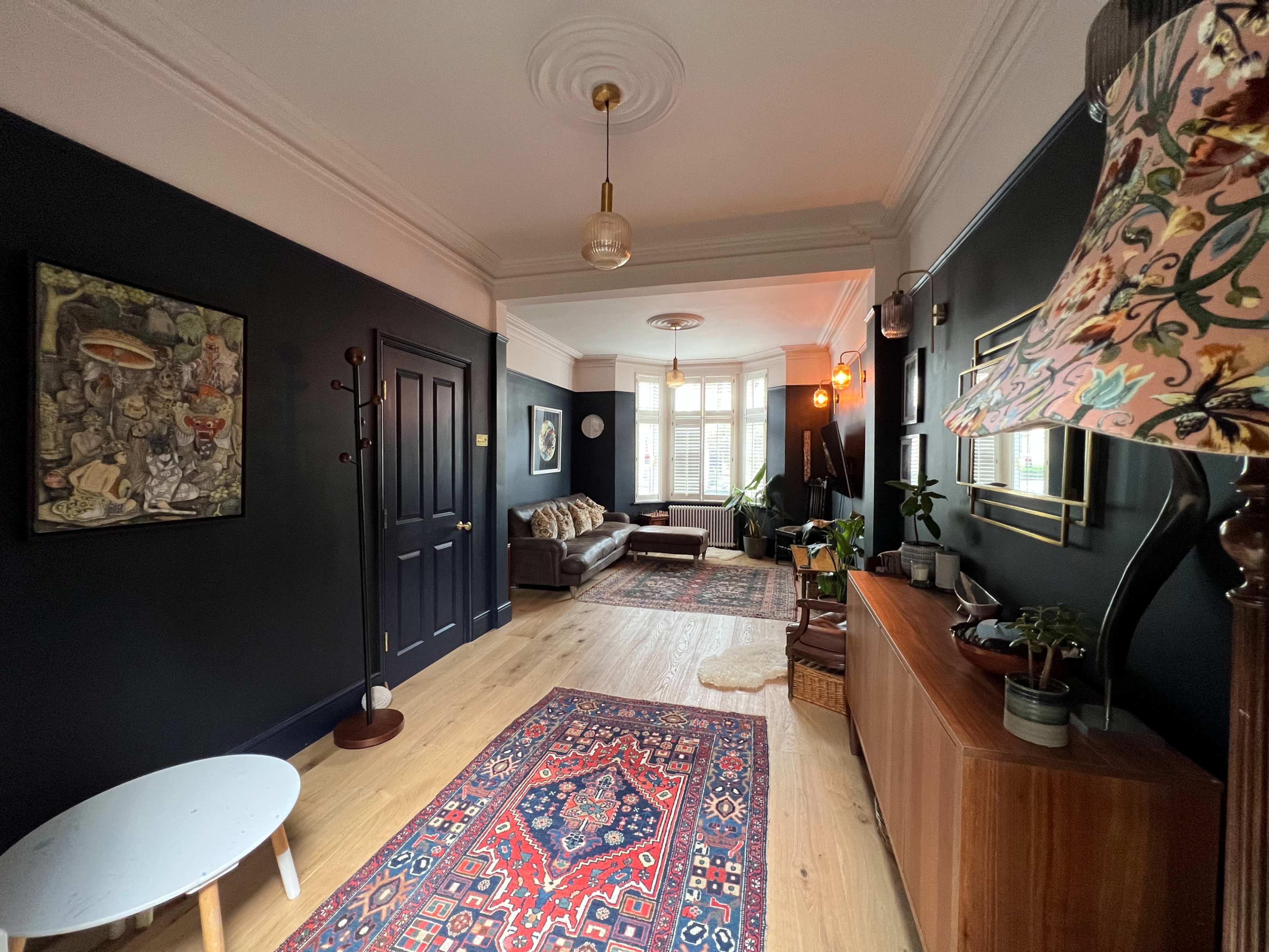 A stylish hallway with black walls, a wooden floor, a vintage rug, and furniture including a sofa, a side table, and a cabinet, all illuminated by pendant lights.
