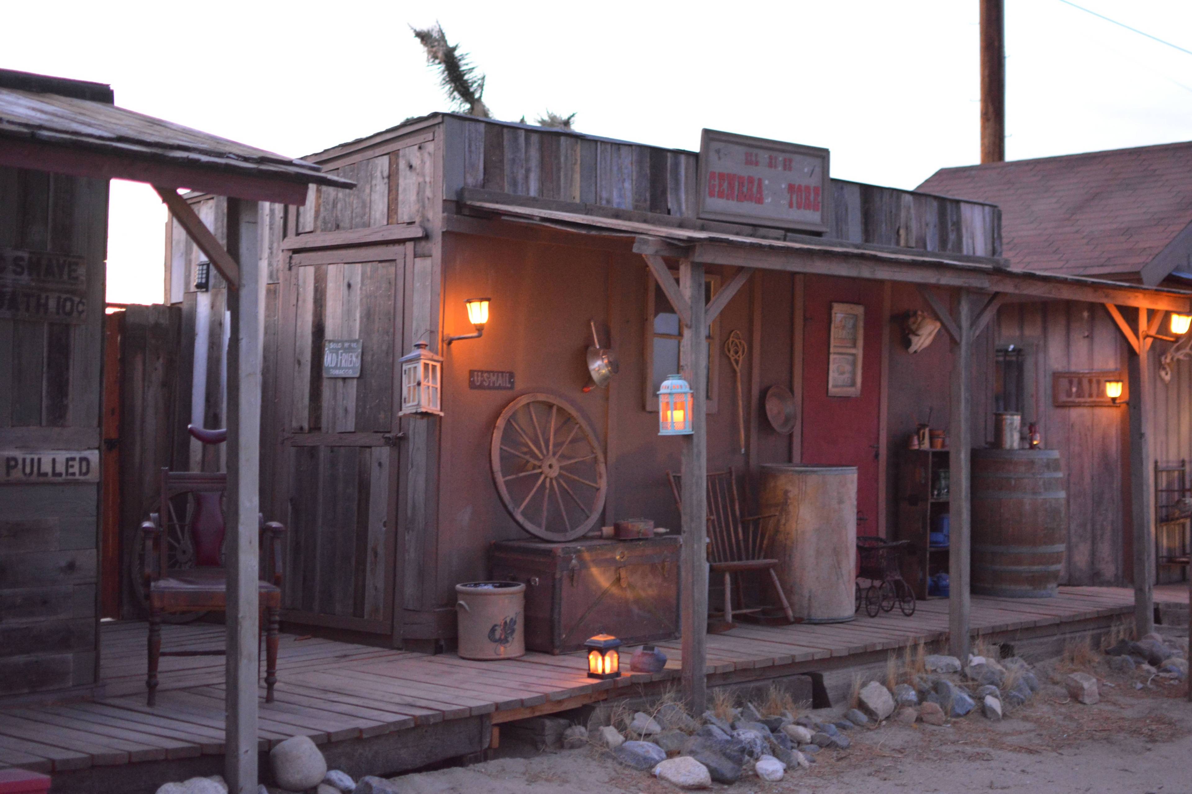 A rustic Western-style storefront with wooden structures, lanterns, and various period-appropriate props along a sandy path.
