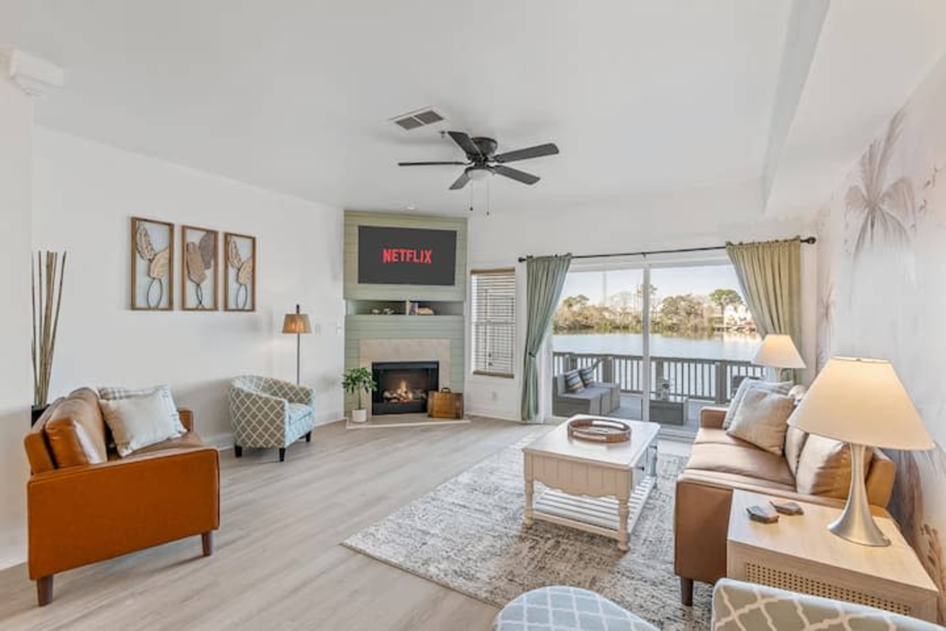 The living room features a cozy arrangement with a television mounted over a fireplace, large windows overlooking a body of water, and various seating options.