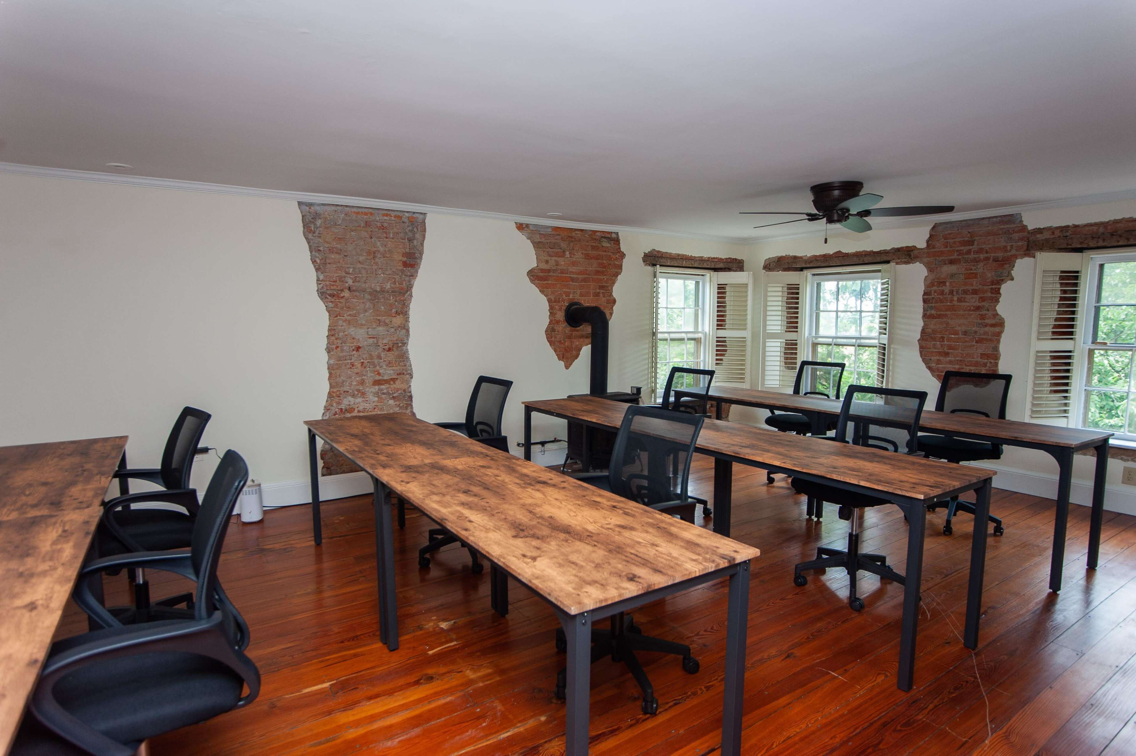 A spacious room with several wooden tables and chairs arranged around the perimeter, exposed brick walls, and large windows.