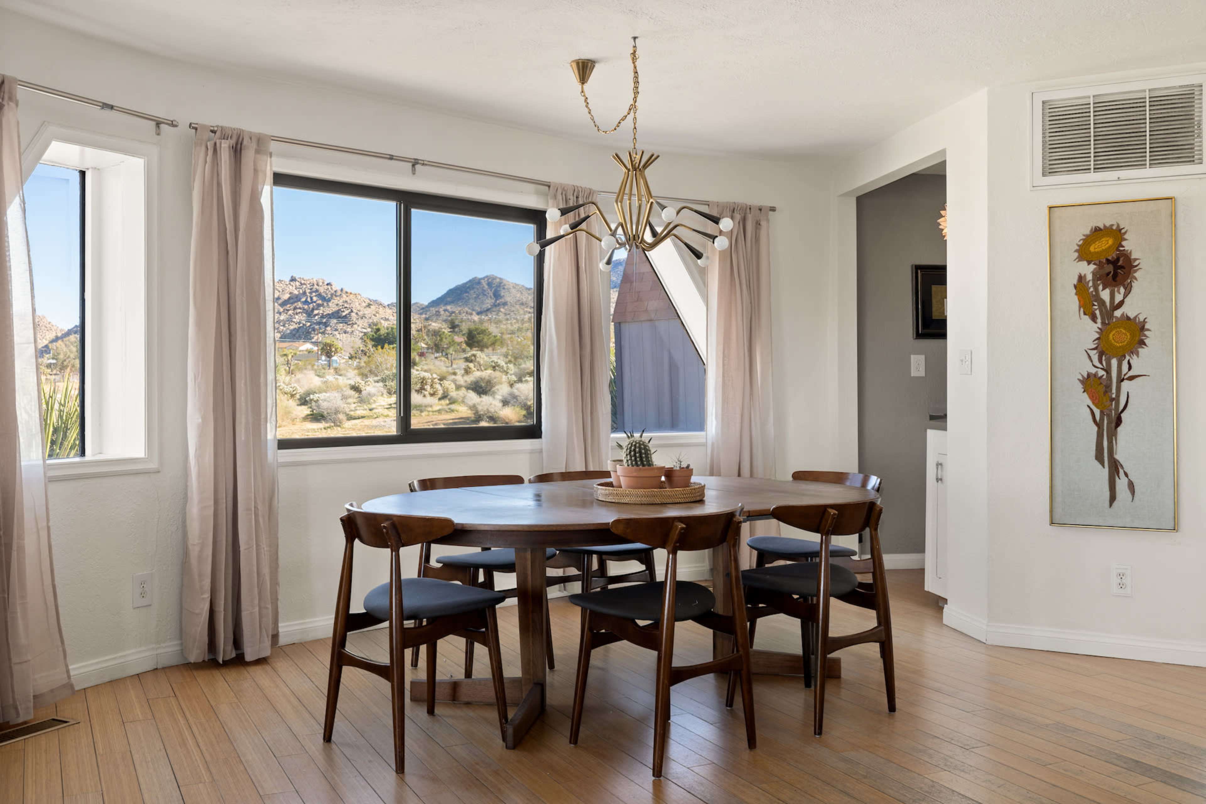 A round wooden dining table with black chairs is situated in a well-lit room featuring large windows that provide a view of mountainous terrain outside.