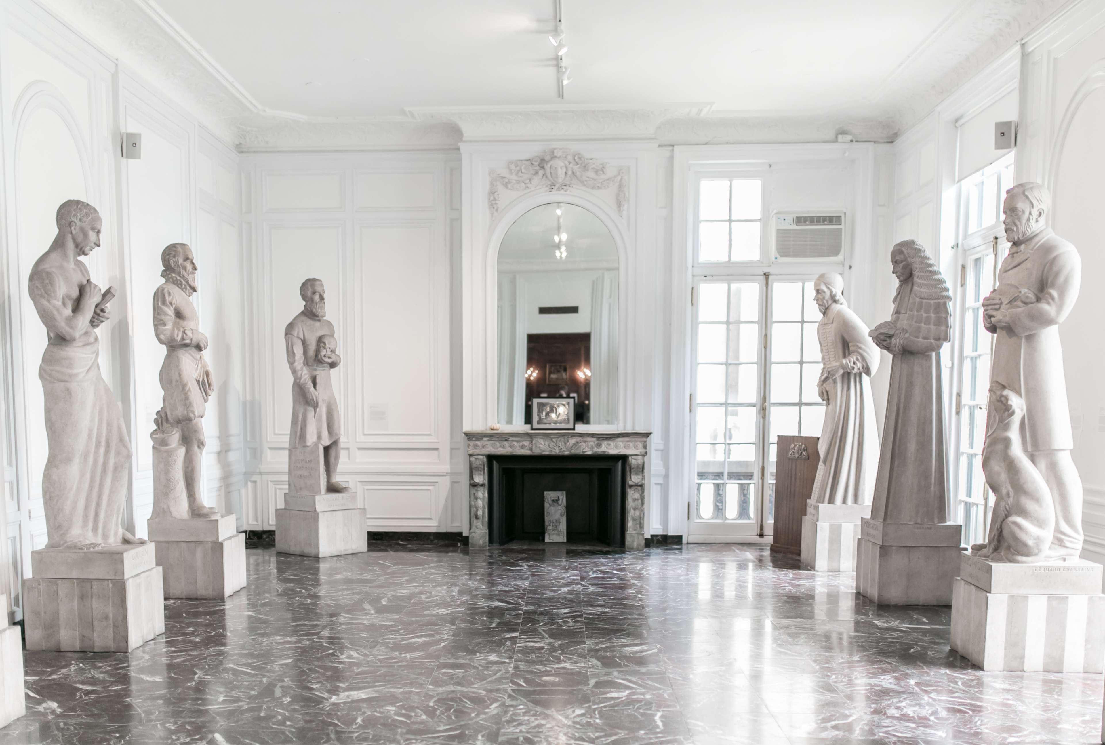 A spacious gallery features six marble statues on pedestals, arranged symmetrically along the walls, with large windows and a mirror reflecting the room's bright, minimalist design.