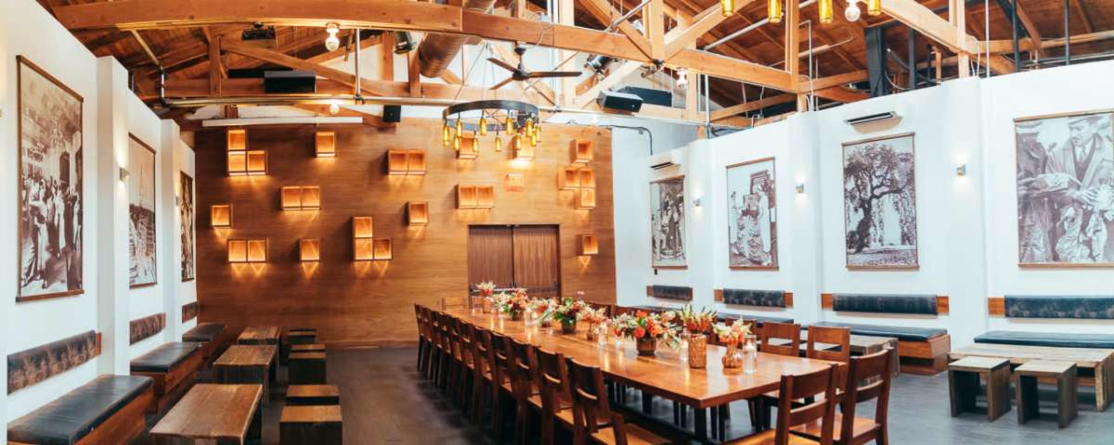 The image shows a spacious dining room with a long wooden table, benches along the walls, and framed photographs hanging on the white walls under a wooden ceiling with exposed beams.