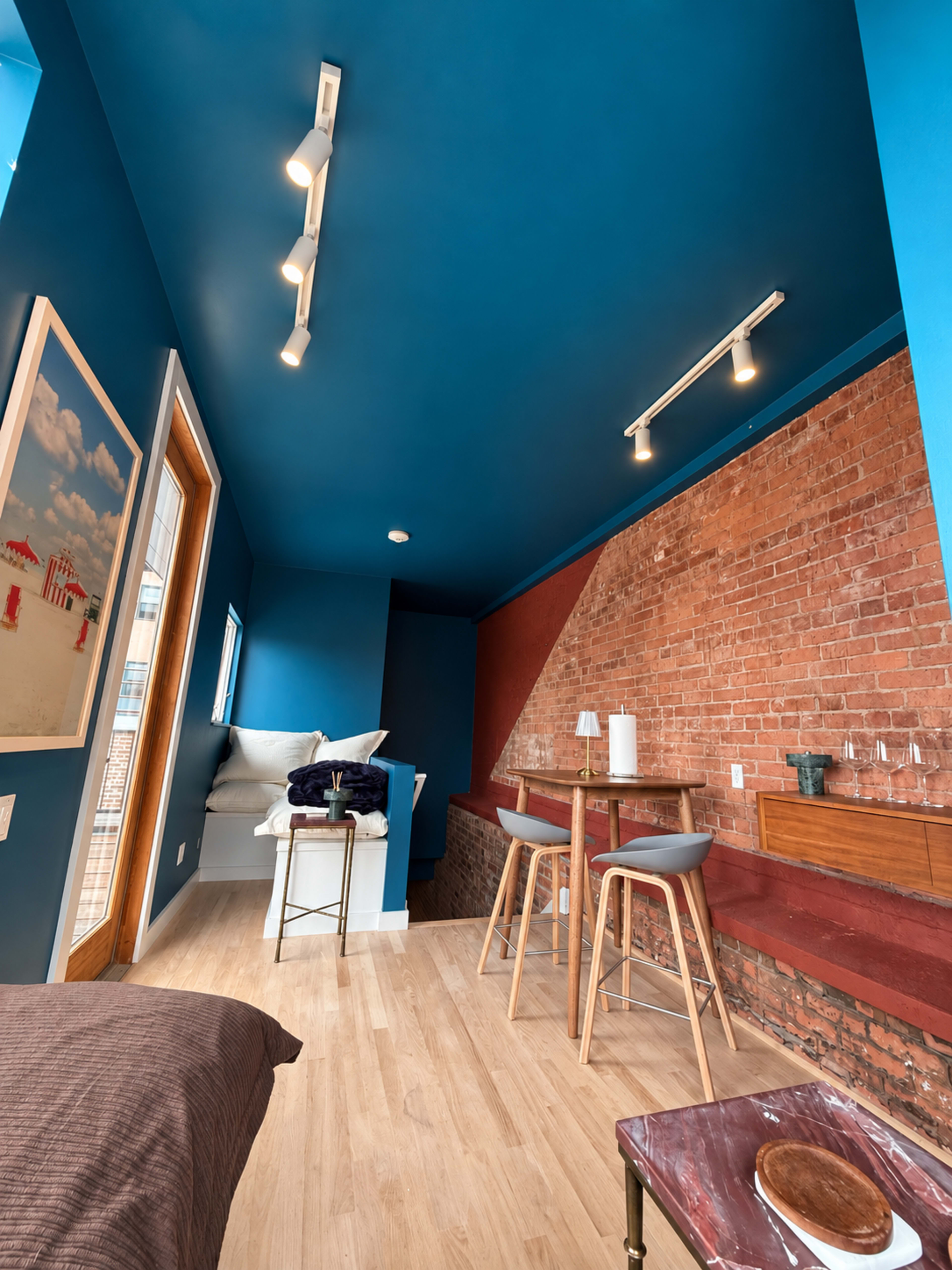 The image shows an interior space featuring blue walls, exposed brick, and a small dining table with stools.