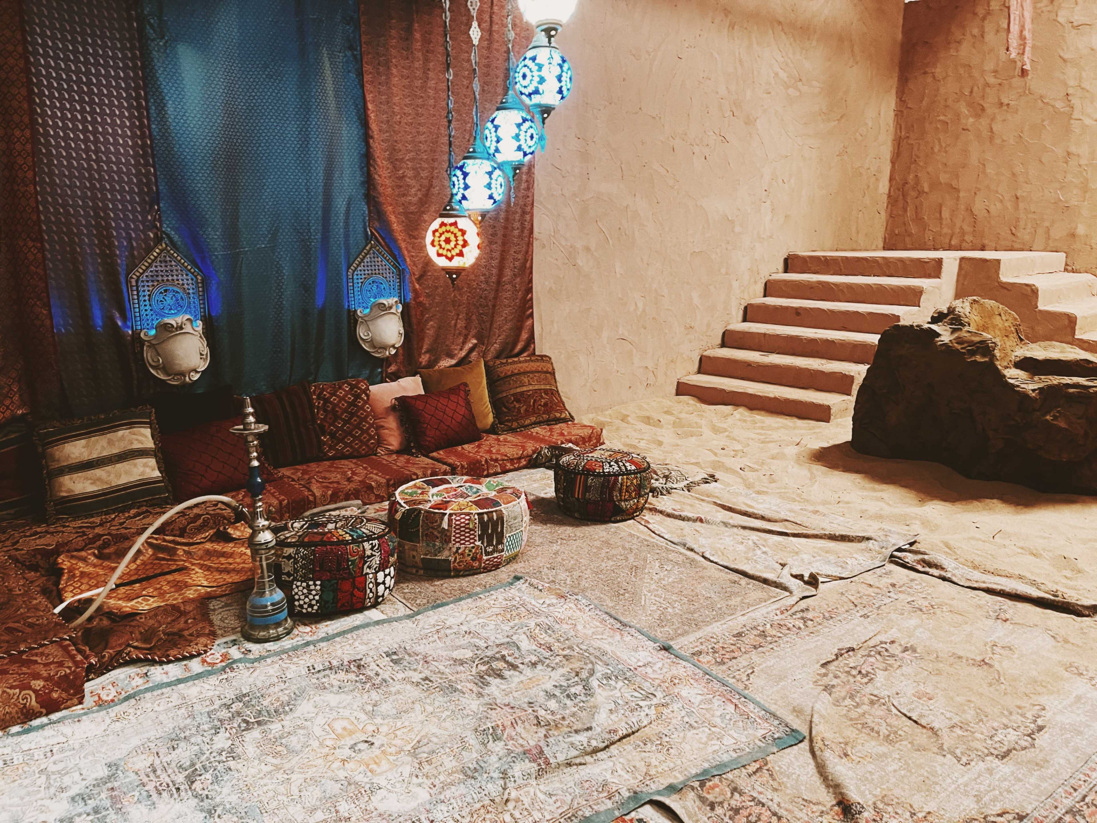 A decorated interior room with colorful cushions and rugs, ornate lamps hanging from the ceiling, and a staircase leading to a different level.