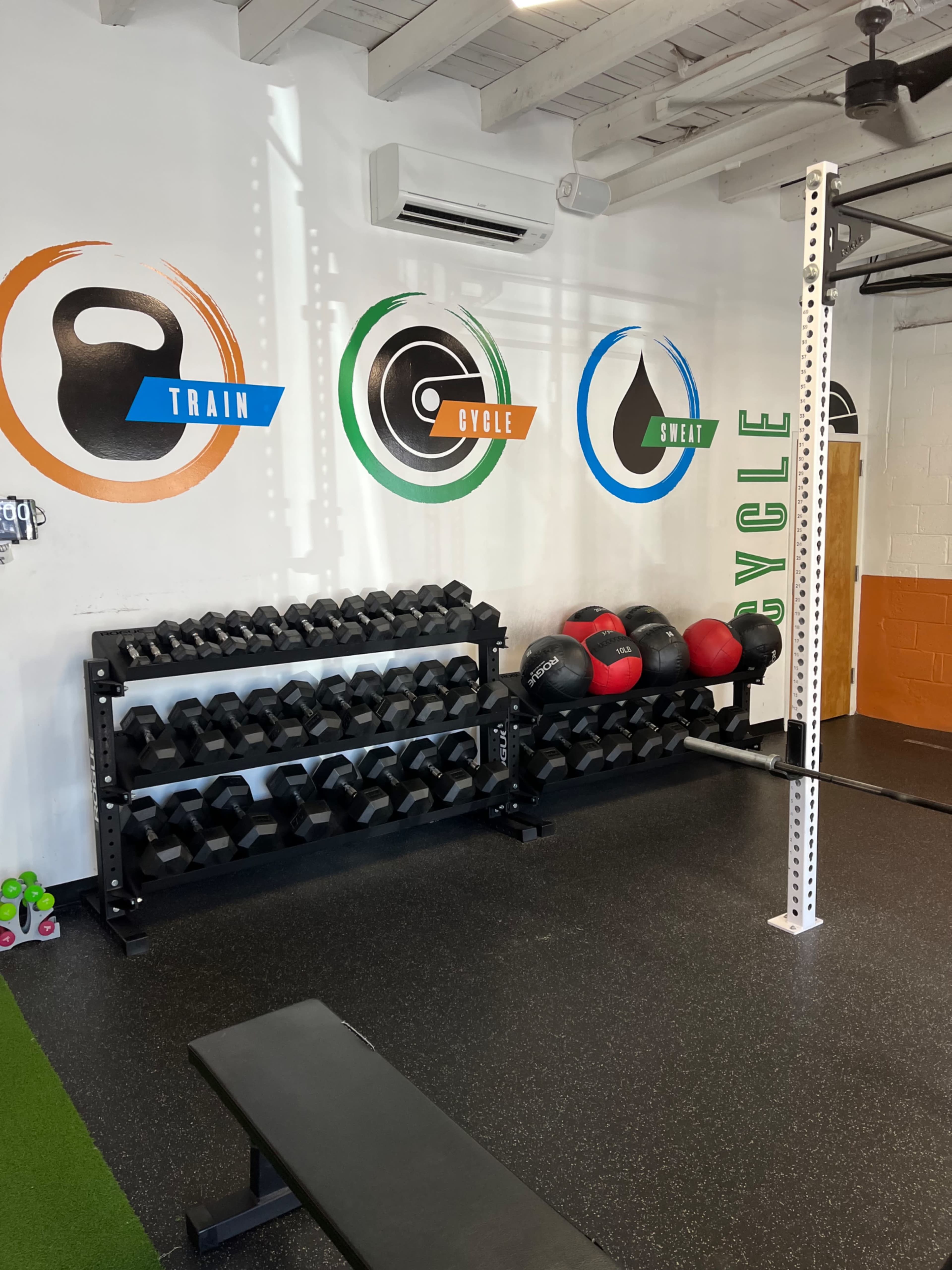 A gym interior featuring a rack of dumbbells, medicine balls, and a bench against a wall with fitness-themed graphics.
