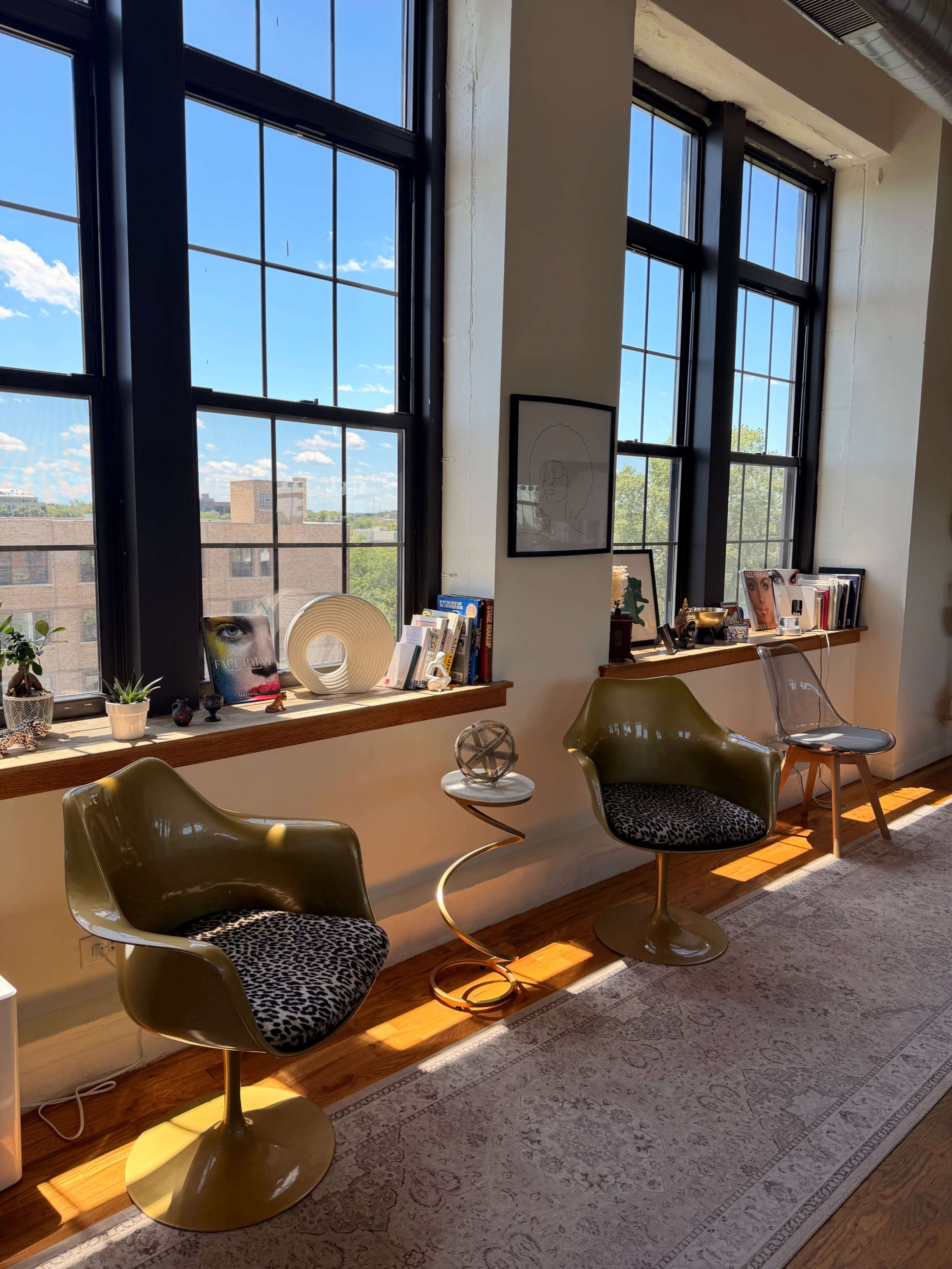 The image shows a bright room with two green chairs, a small side table, and large windows letting in natural light, adorned with books and decorative items on the windowsill.