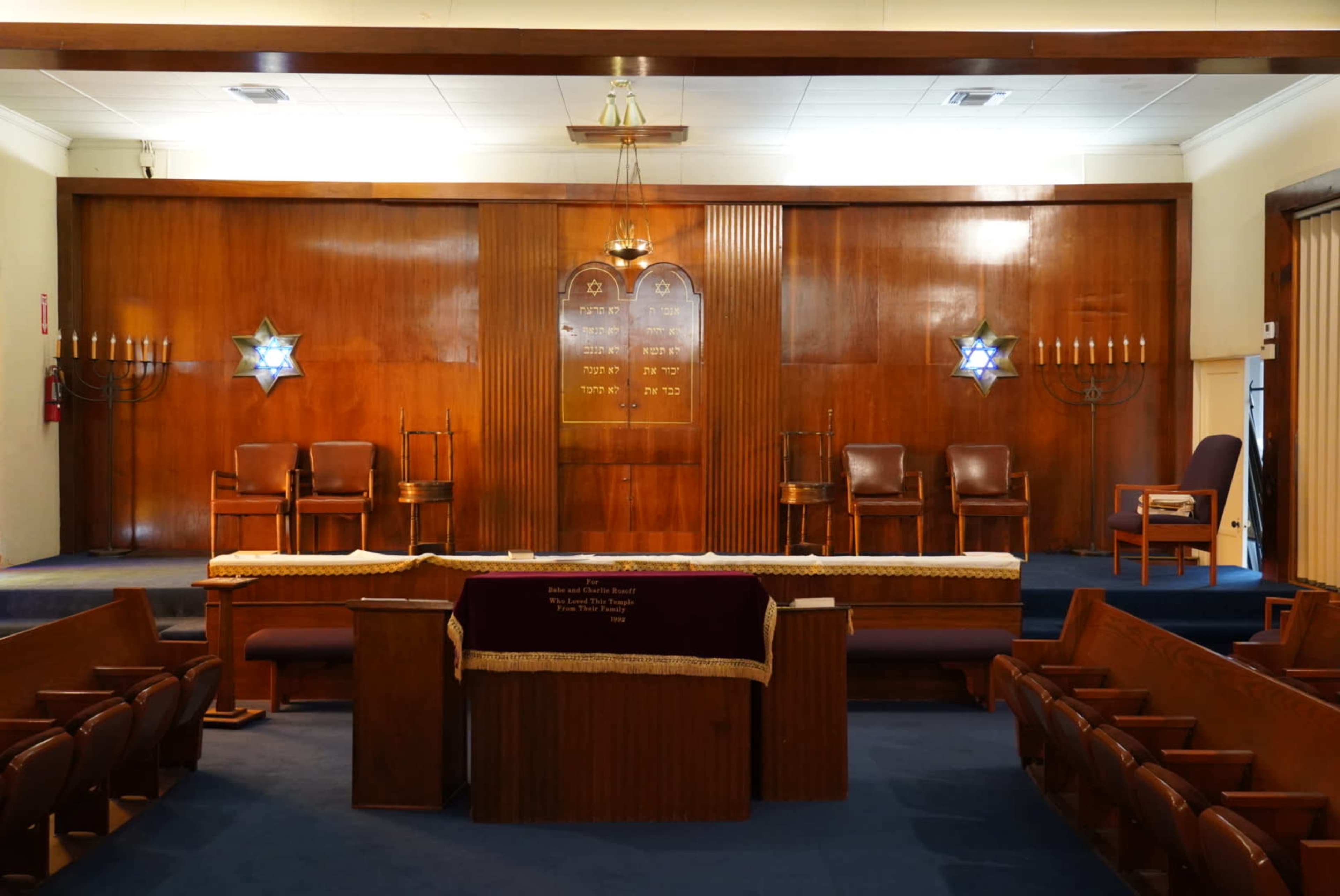 The interior of a synagogue features wooden paneling, a raised bimah with chairs, and decorative elements like a Star of David on the walls.