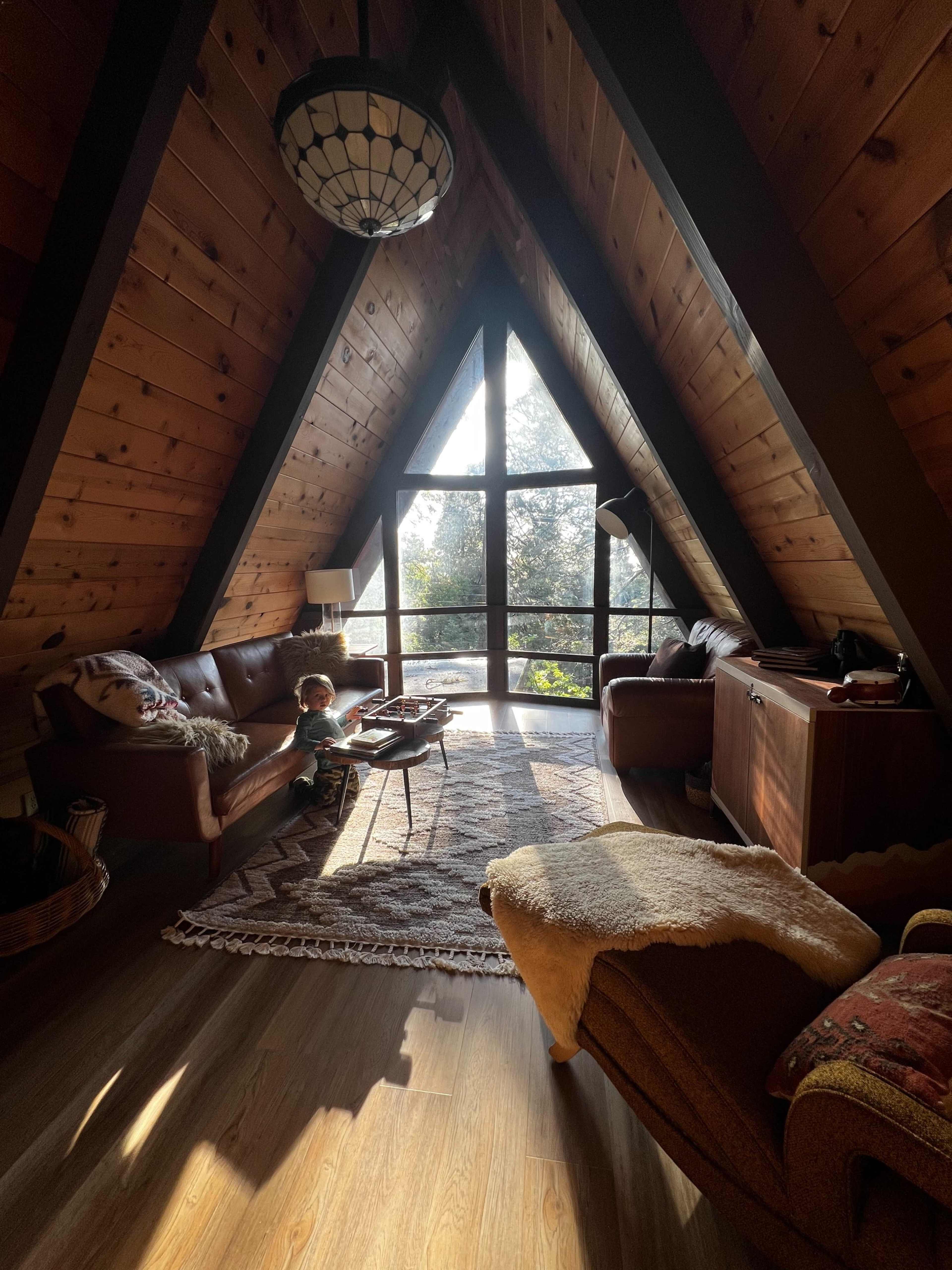 A child stands next to a coffee table in a cozy A-frame living room filled with natural light from large windows.