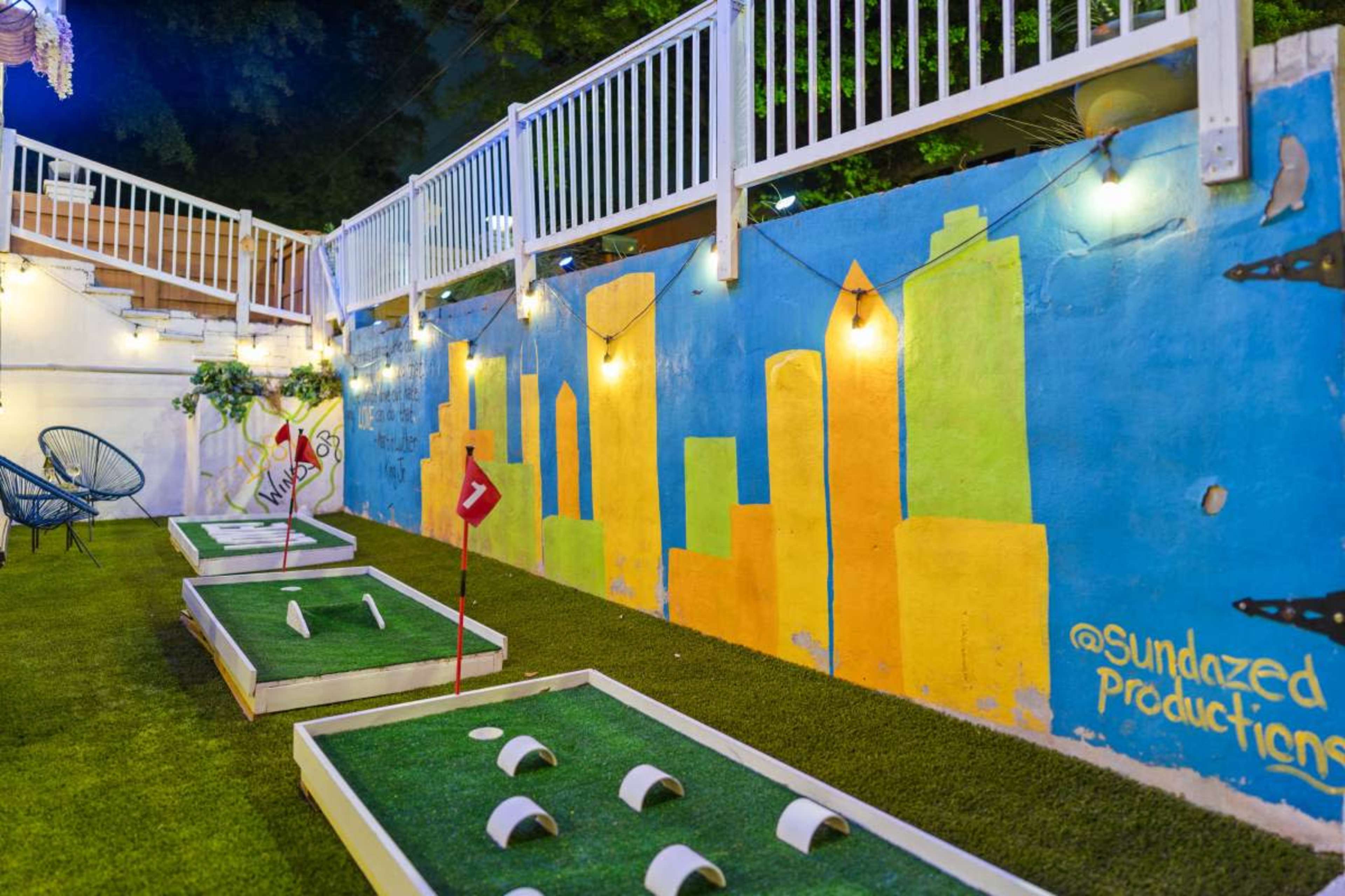 The image shows a vibrant outdoor space featuring several white, raised platforms for a game, set against a brightly painted mural of city skyline silhouettes on a wall.