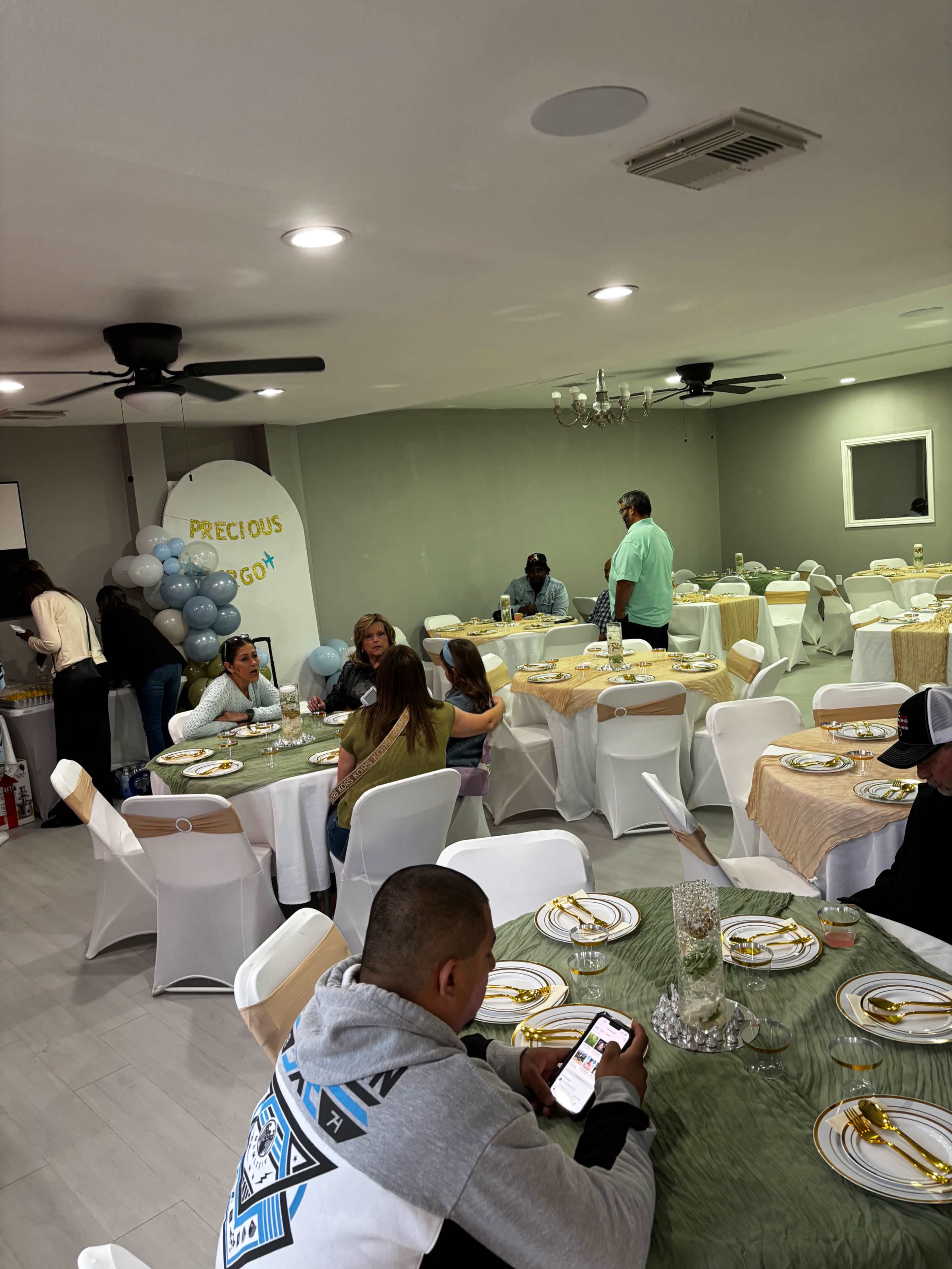 The image shows a banquet hall with neatly arranged tables set for an event, featuring decorative tablecloths, plates, and balloons, while guests are mingling around.