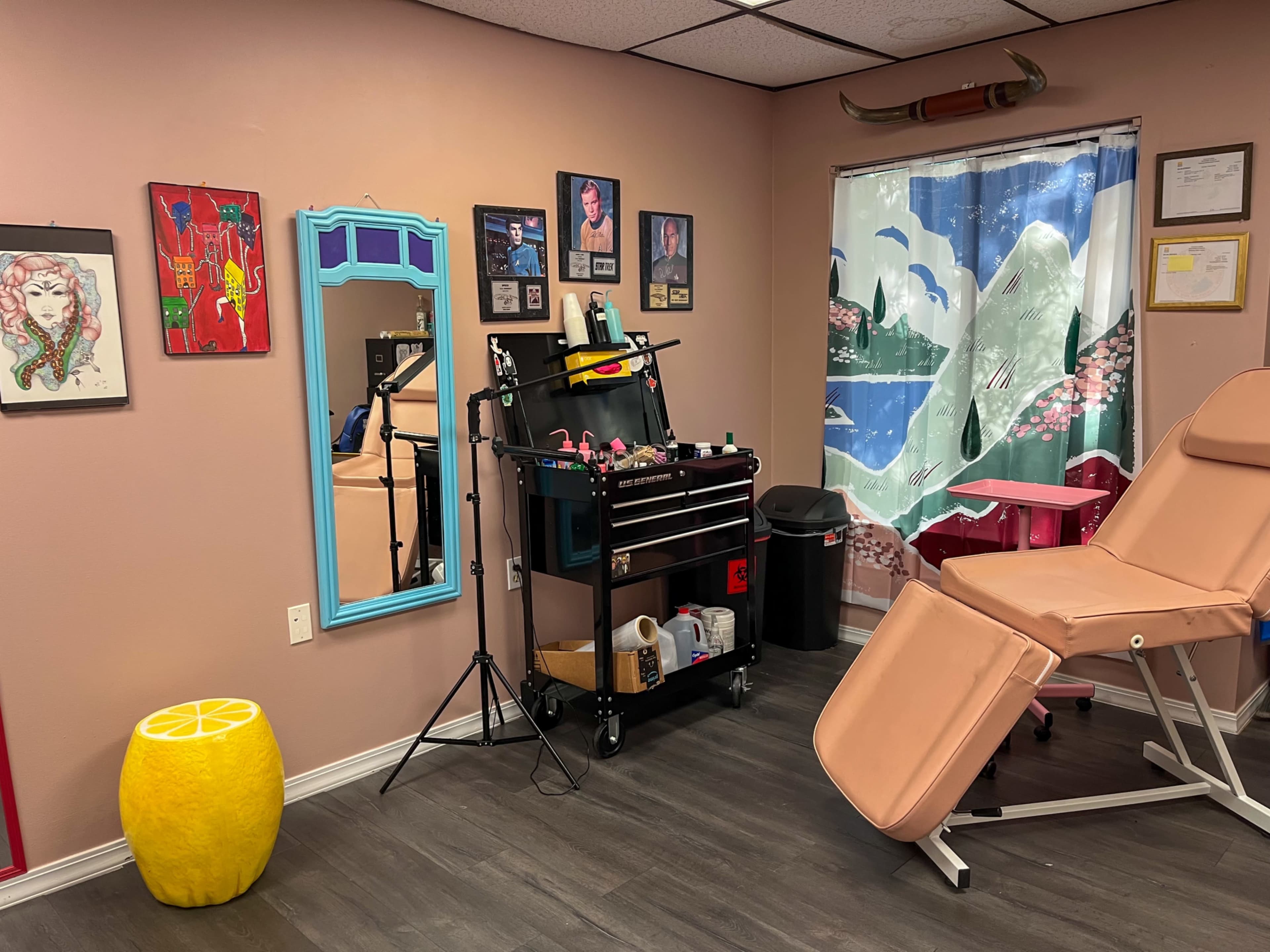 The image shows a tattoo studio with a pink treatment chair, a tool cart filled with equipment, a large window curtain depicting a mountain scene, and various artworks on the walls.