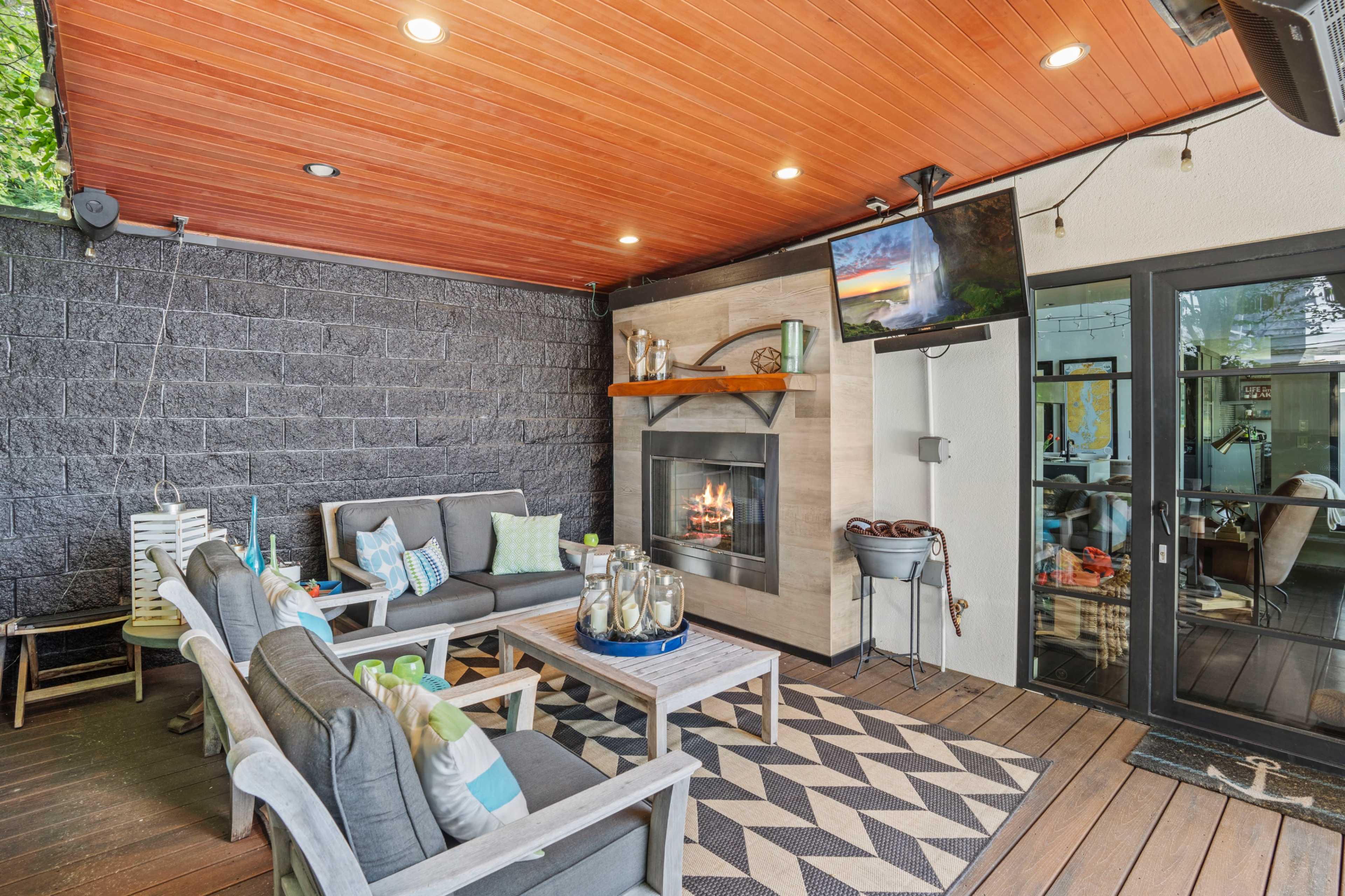 A cozy outdoor seating area with comfortable gray sofas, a coffee table, a fireplace, and a mounted television under a wooden ceiling.