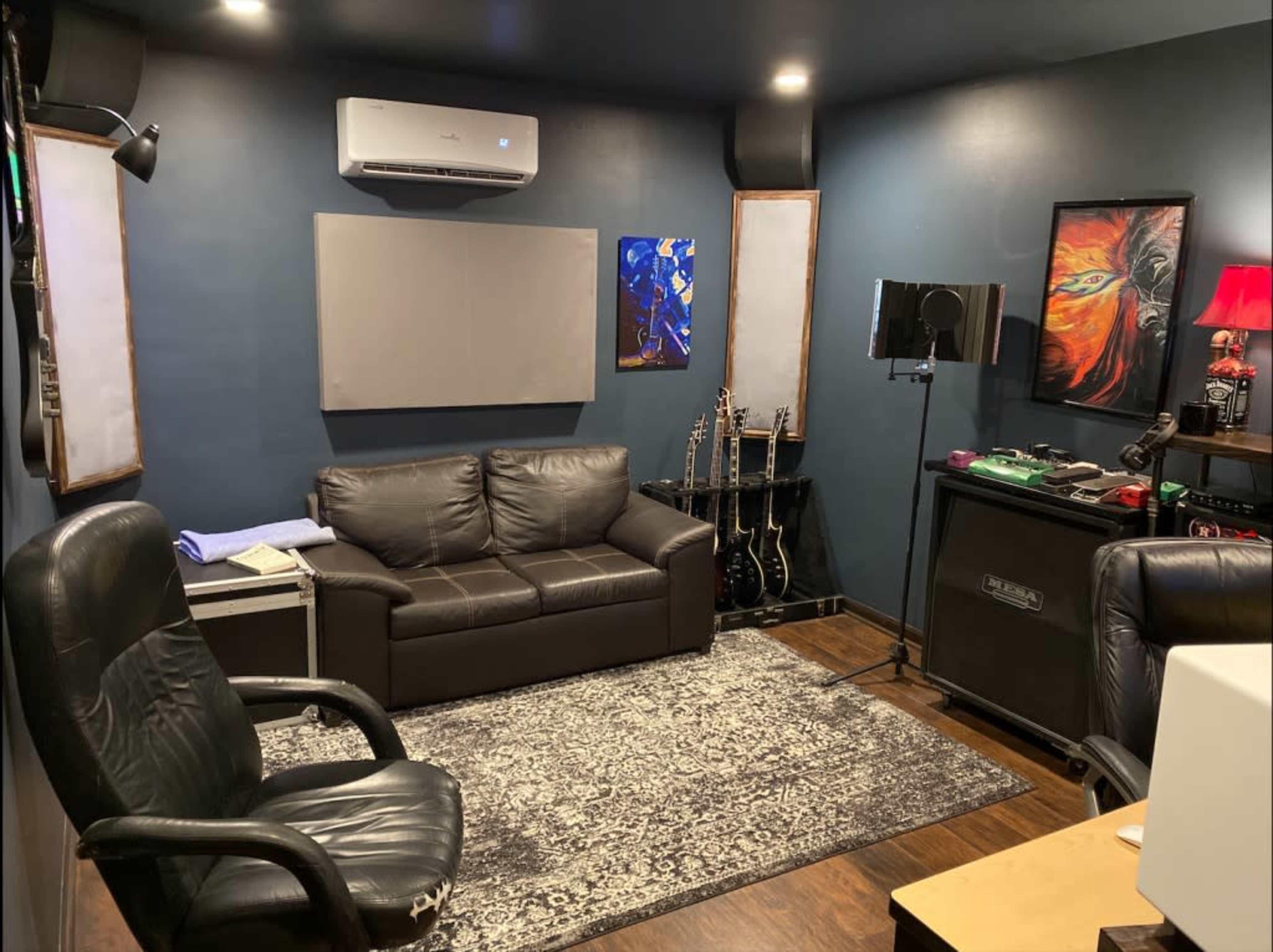 A cozy music studio featuring a leather sofa, a coffee table, two guitars, an amplifier, and acoustic panels on the walls.