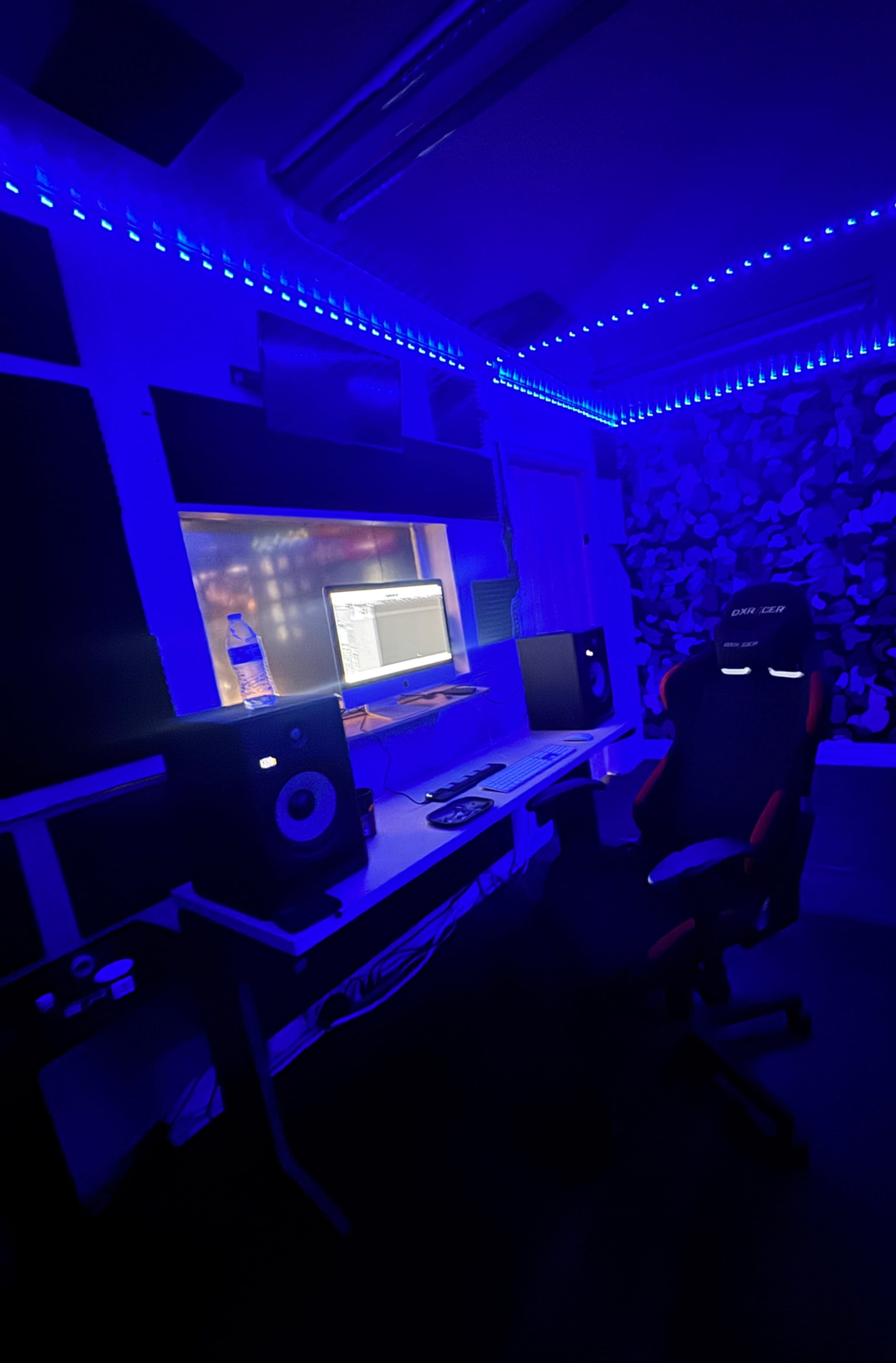The image shows a dimly lit music production studio with blue LED lights, a computer screen, studio monitors, and an ergonomic chair.