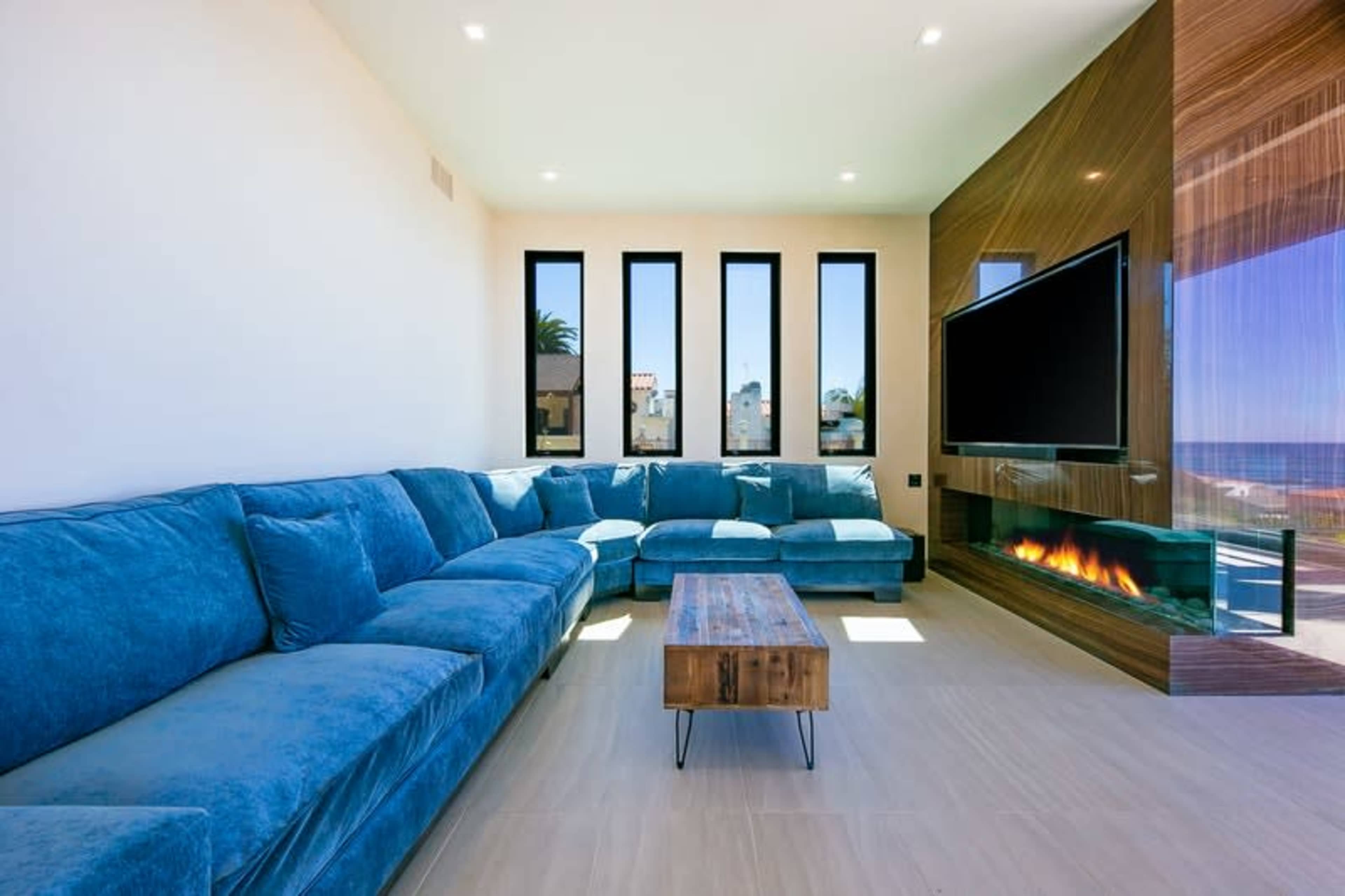 A modern living room features a large blue sectional sofa, a wooden coffee table, and a wall-mounted television above a sleek fireplace, with large windows revealing an outdoor view.