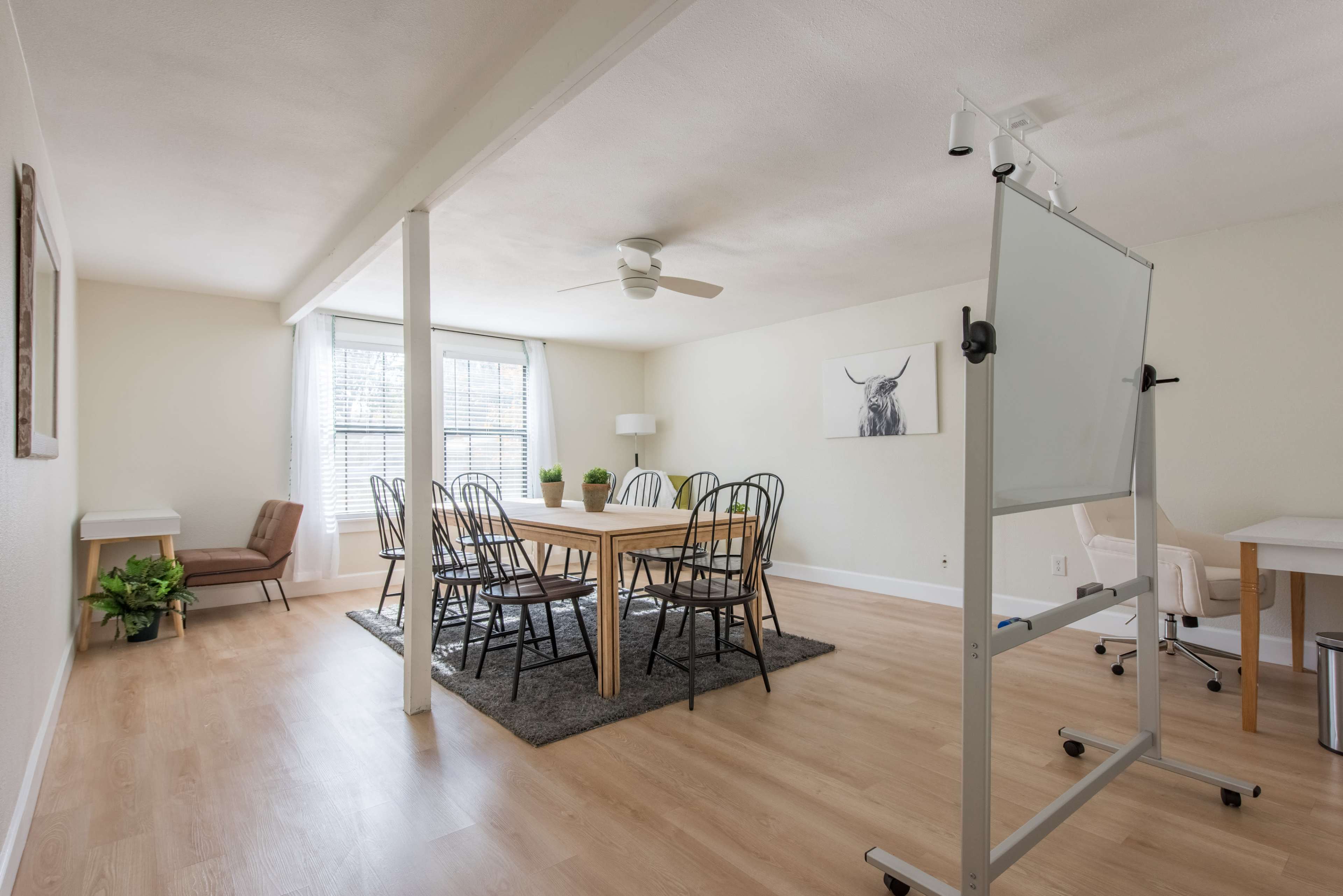 A light-filled dining area features a wooden table surrounded by black metal chairs, with a whiteboard and a comfortable seating area in the corner.