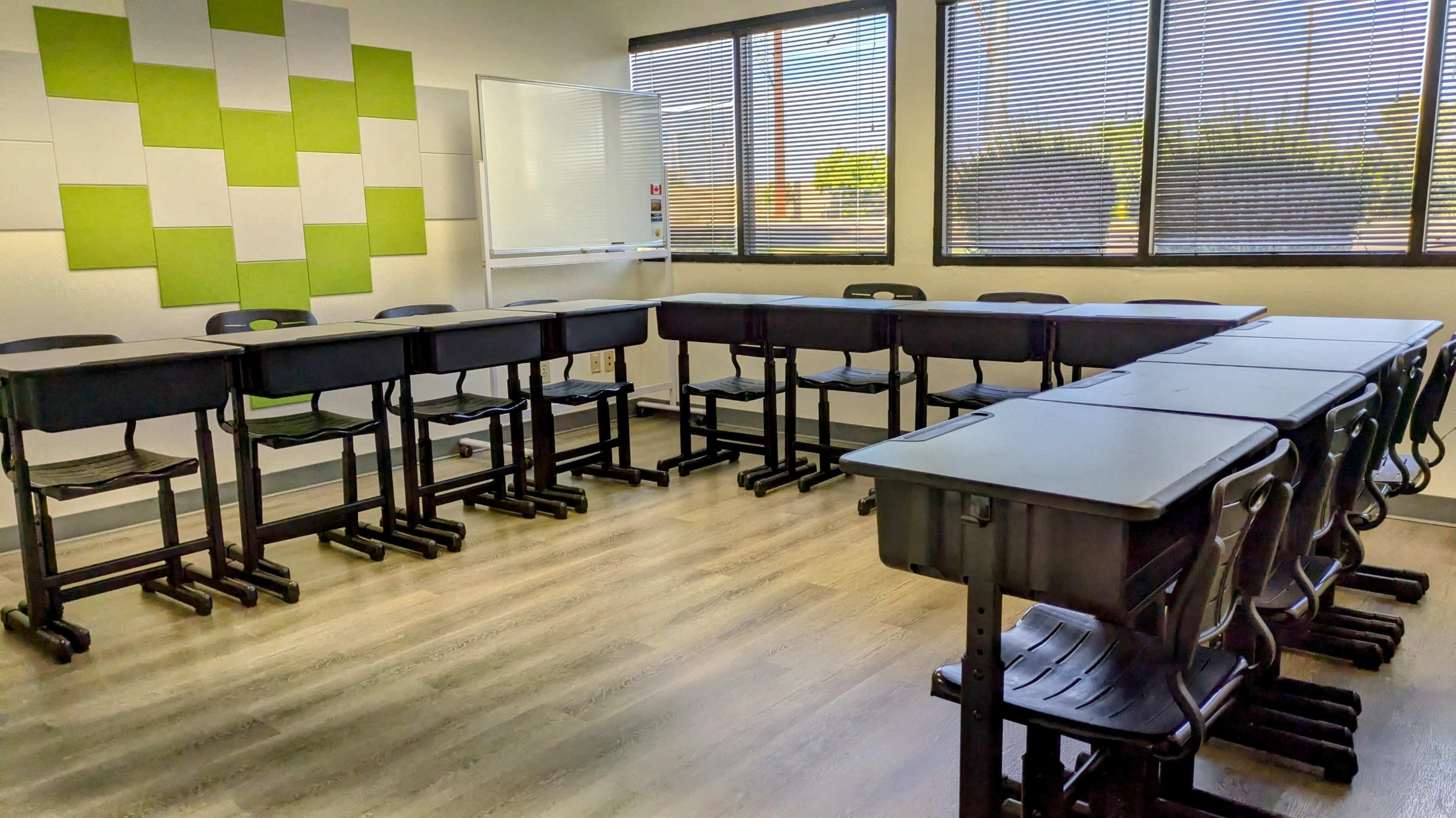 The image shows a classroom arranged with several black desks and chairs in a U-shape, surrounded by large windows and accented by a green and white wall design.