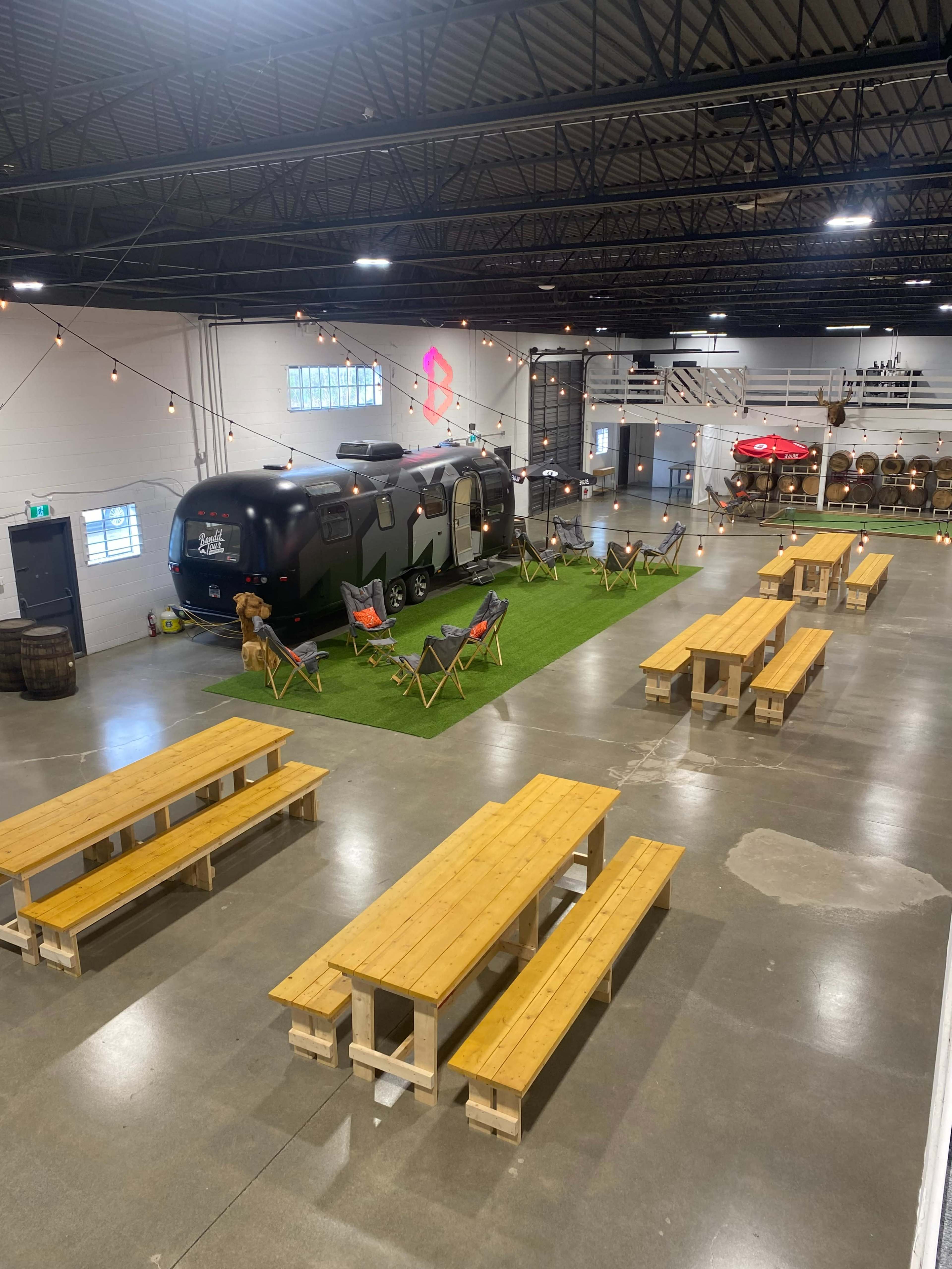 An industrial space with a black Airstream trailer on artificial grass, surrounded by wooden picnic tables and lounge chairs.