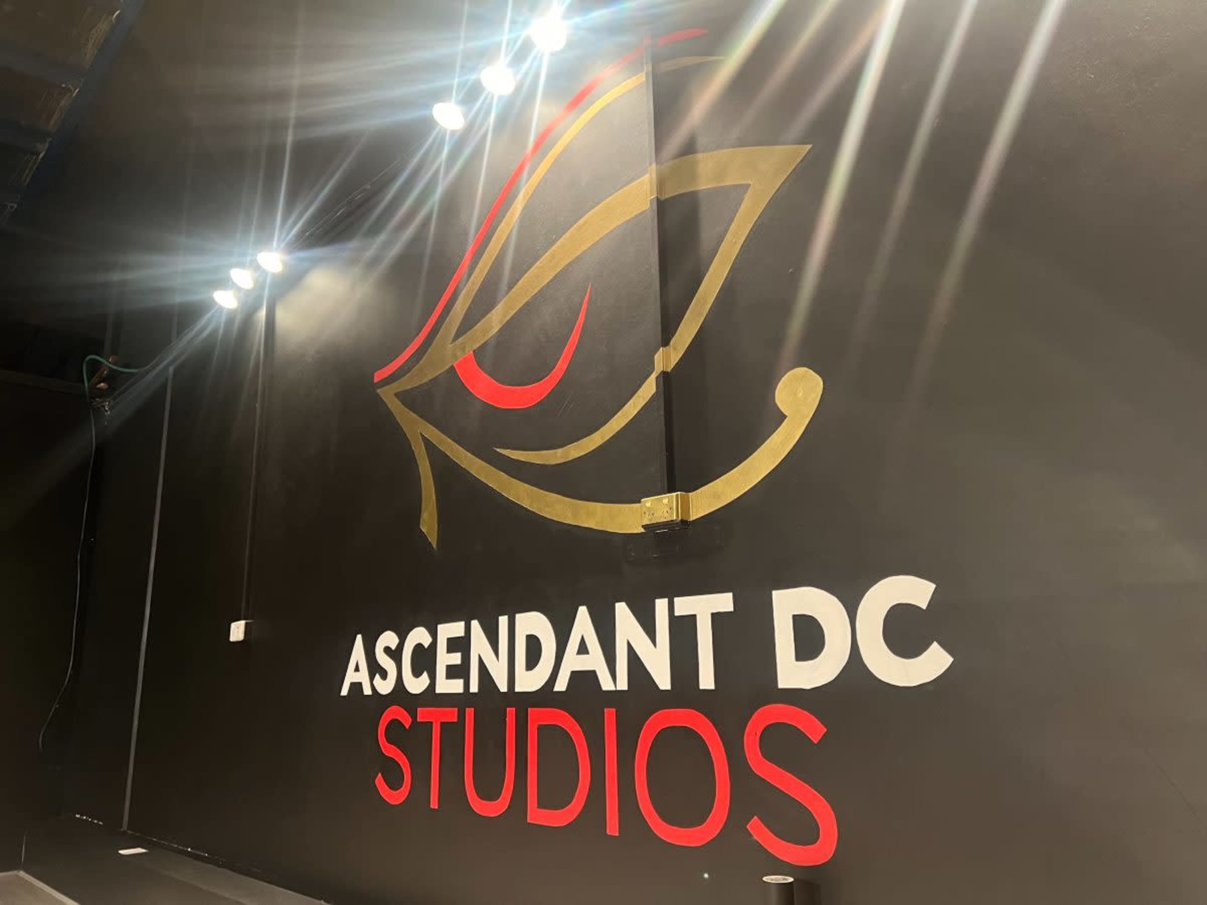 The image shows a black wall featuring the text "ASCENDANT DC STUDIOS" in bold white and red letters, along with an artistic design of an eye above it.