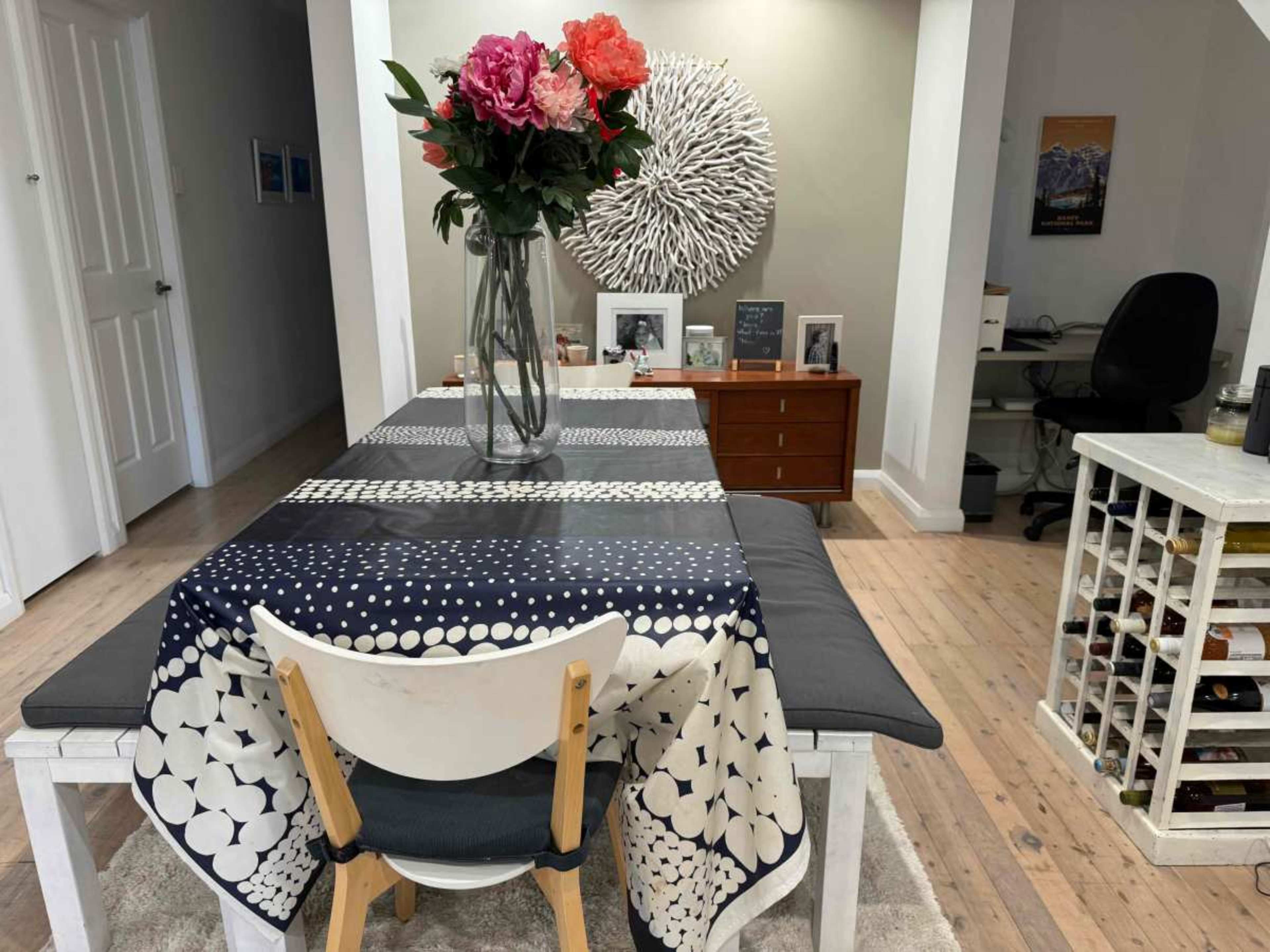 A dining table with a patterned tablecloth is set with a vase of flowers, surrounded by chairs, and there is a sideboard and a workspace visible in the background.