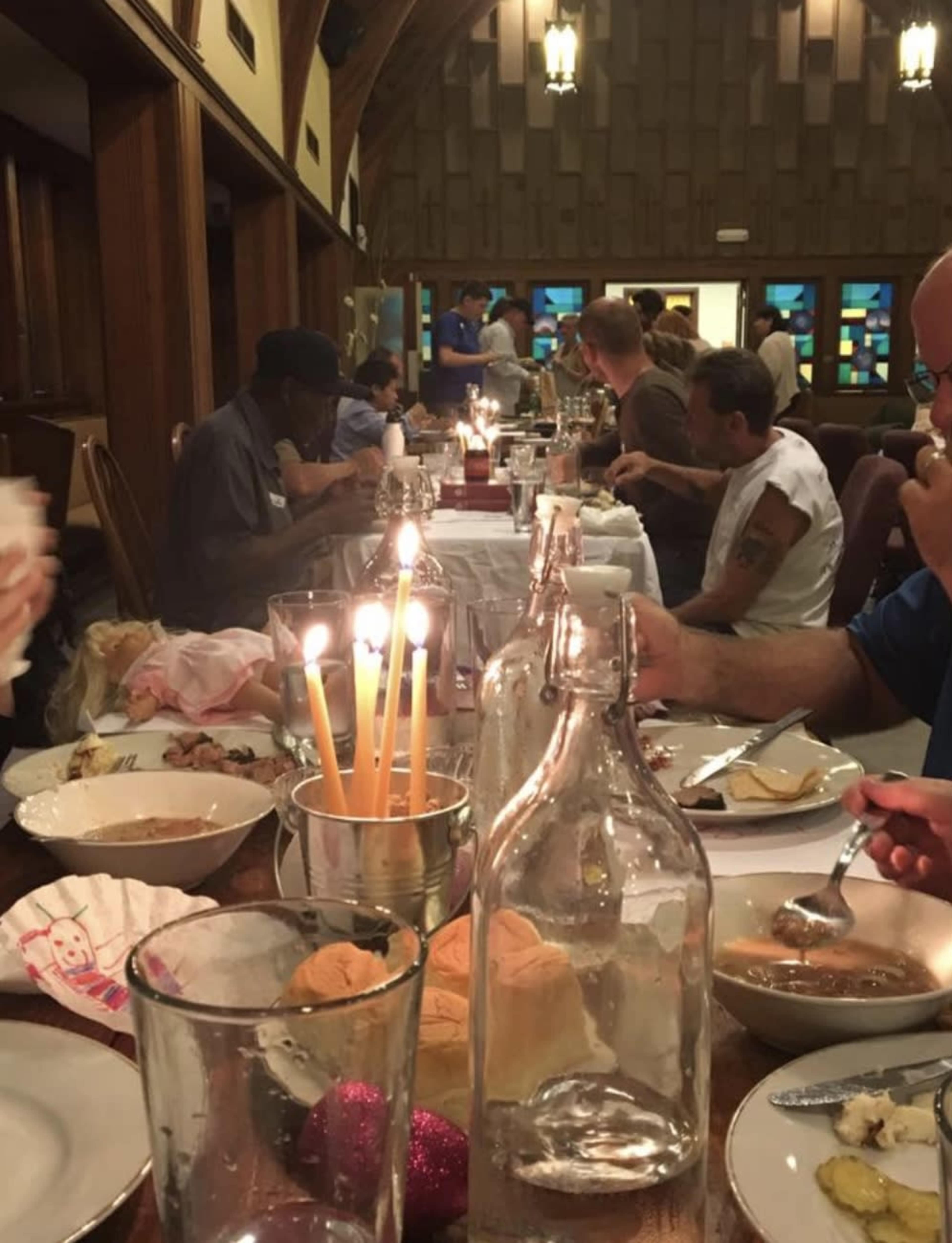 A long dining table is filled with people eating dinner in a warmly lit room, with candles and various food items visible.
