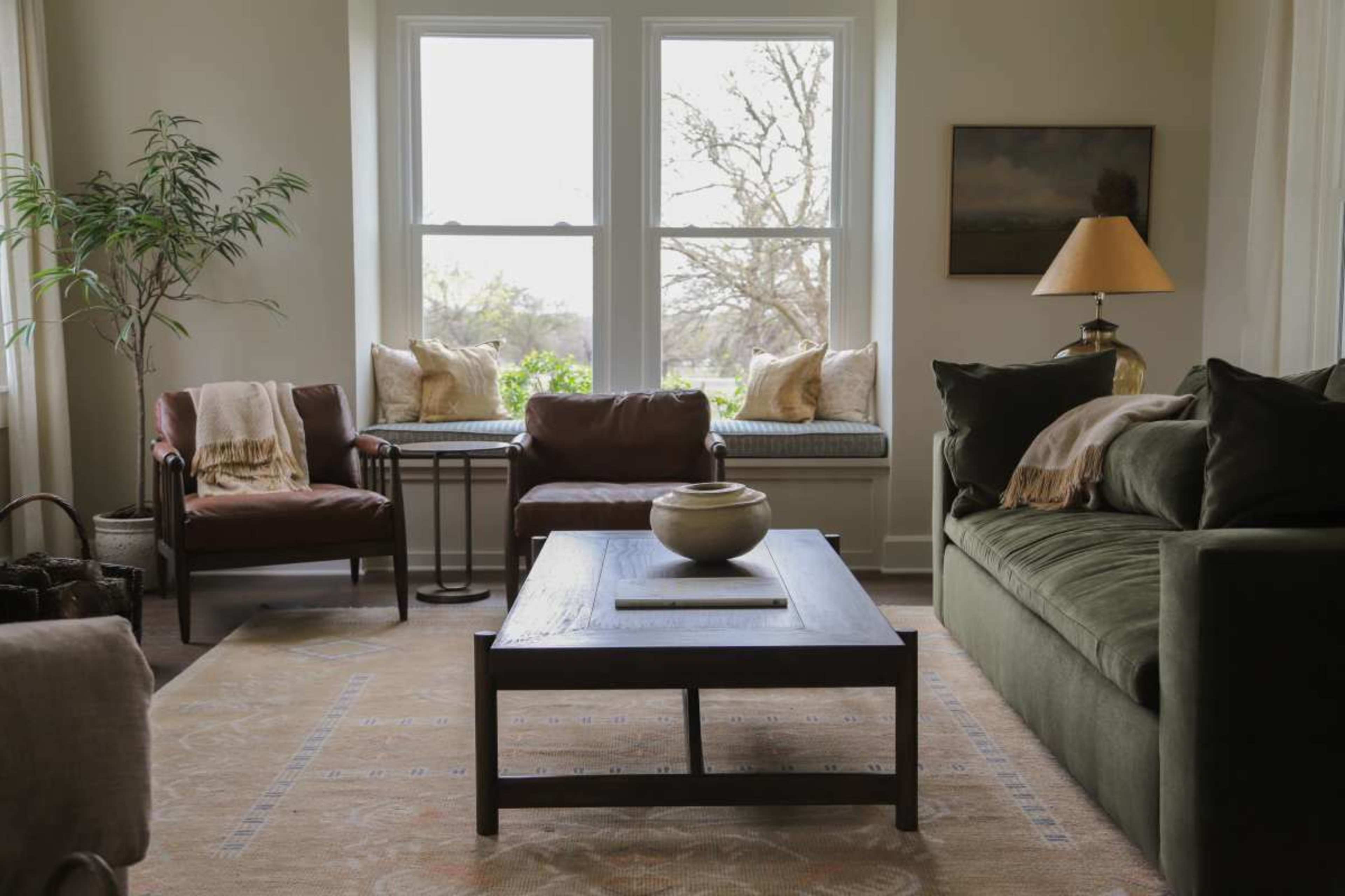 The living room features a green sofa, two brown chairs, a wooden coffee table, and large windows with a view of greenery outside.
