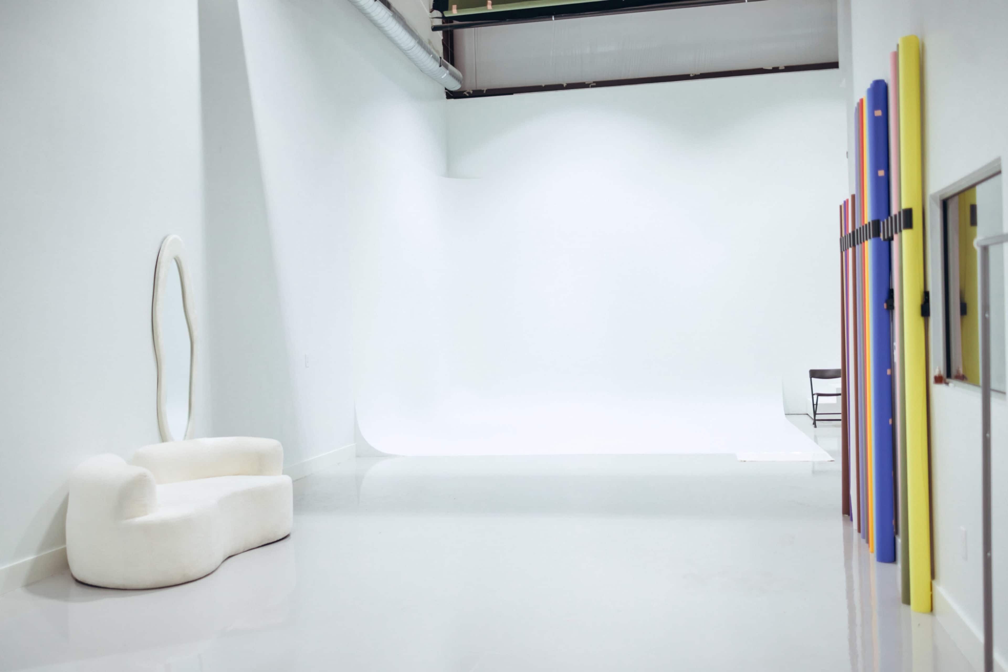The image shows a spacious, minimalistic studio with a white backdrop and a curved white seating area on one side.