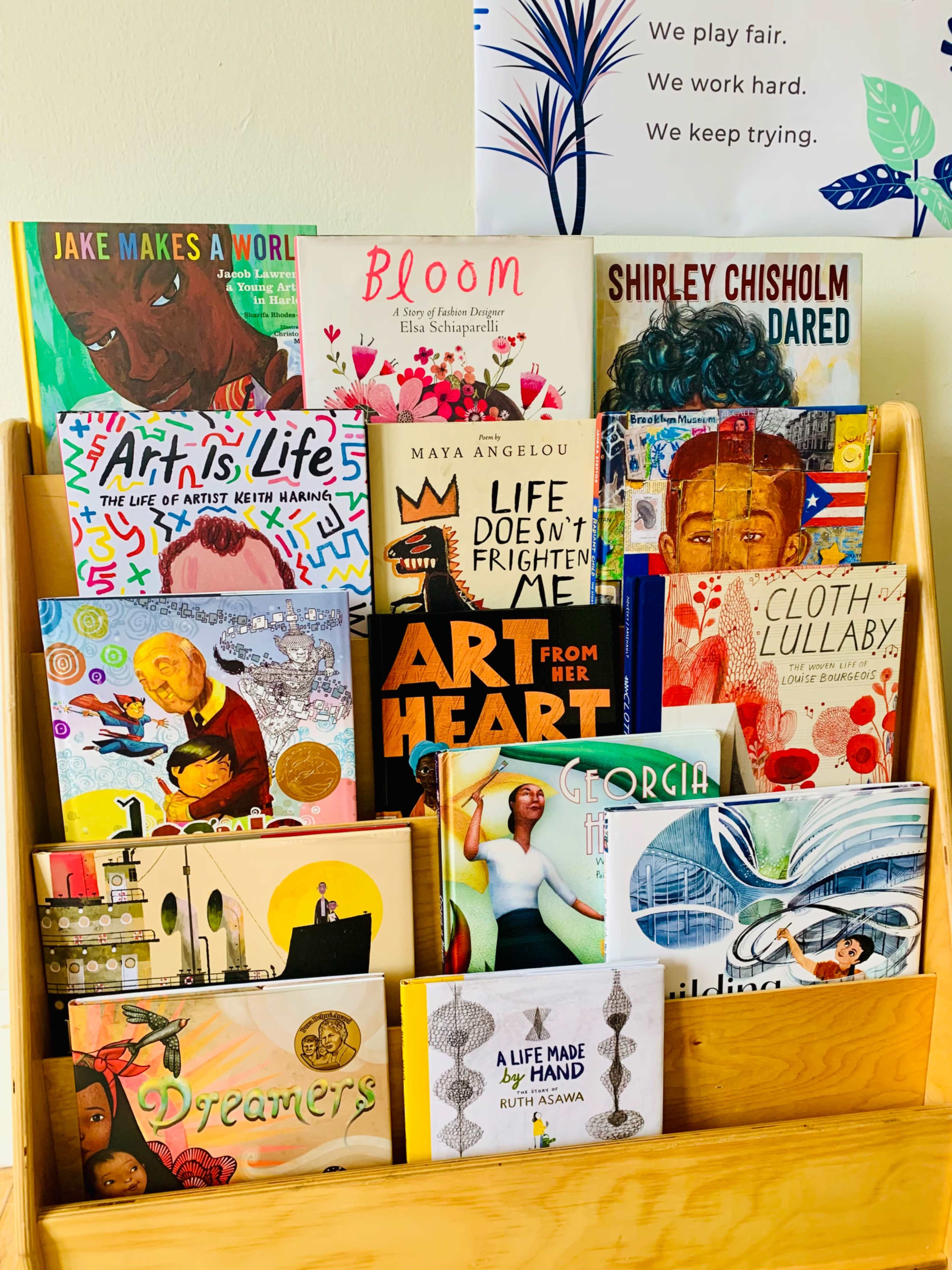 A wooden book display contains various children's books with colorful covers, highlighting themes of art, life, and notable figures.