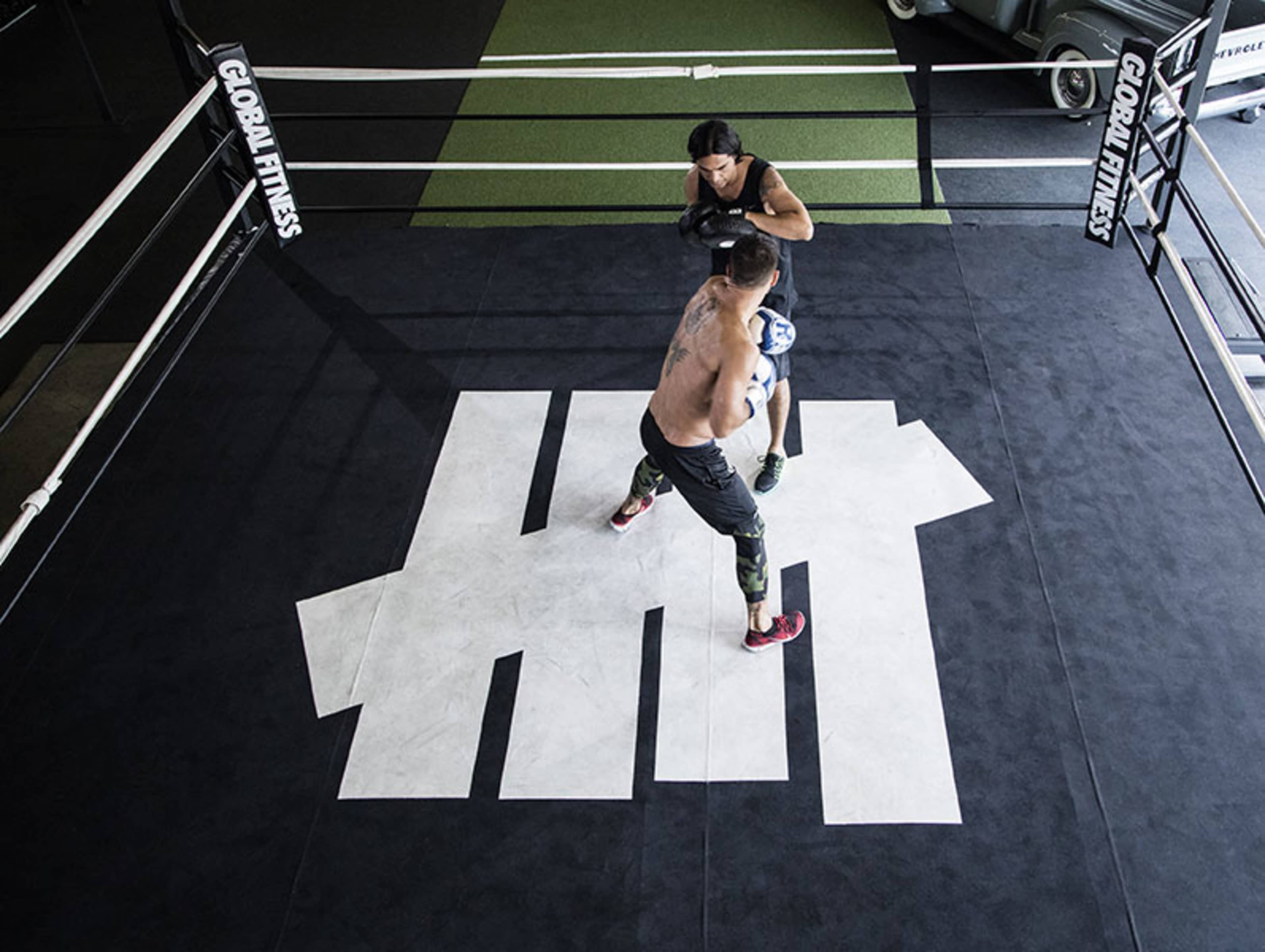 Two individuals are sparring in a boxing ring with the logo of a fitness brand displayed on the floor.