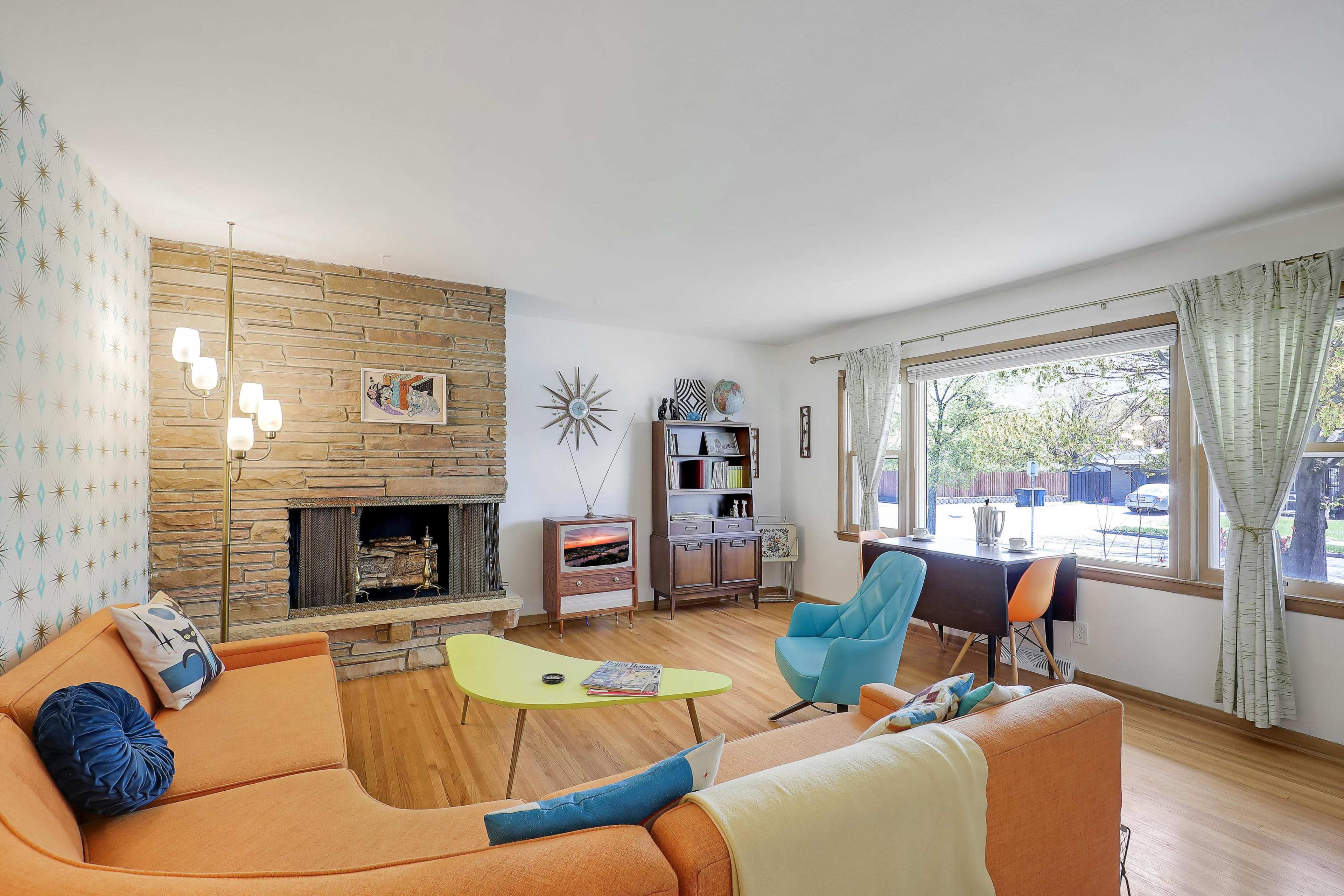 The living room features a stone fireplace, a vibrant orange sofa, a colorful coffee table, and large windows that illuminate the space.