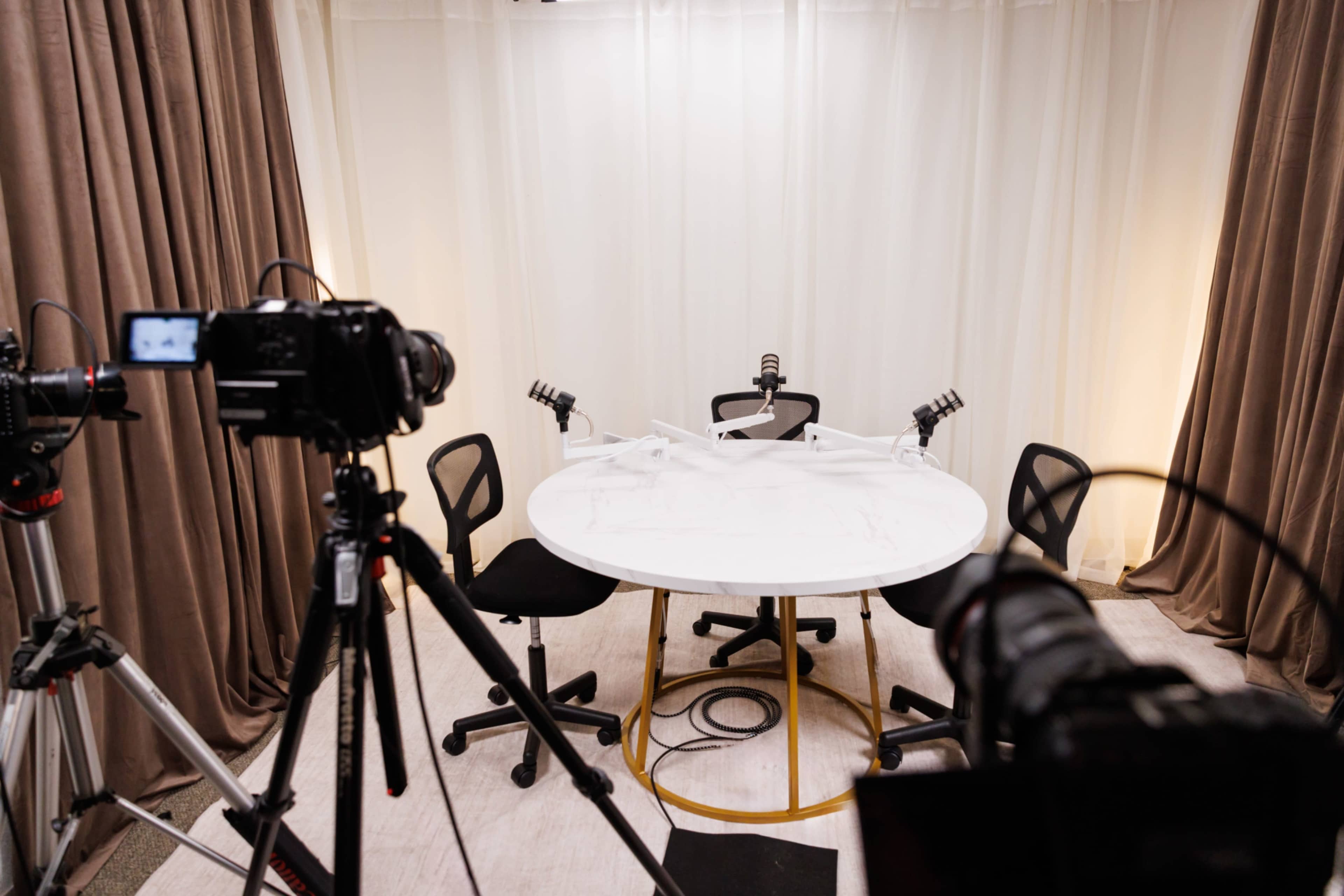 A round marble table surrounded by black chairs is set up under soft lighting in a studio space, with cameras positioned in front.