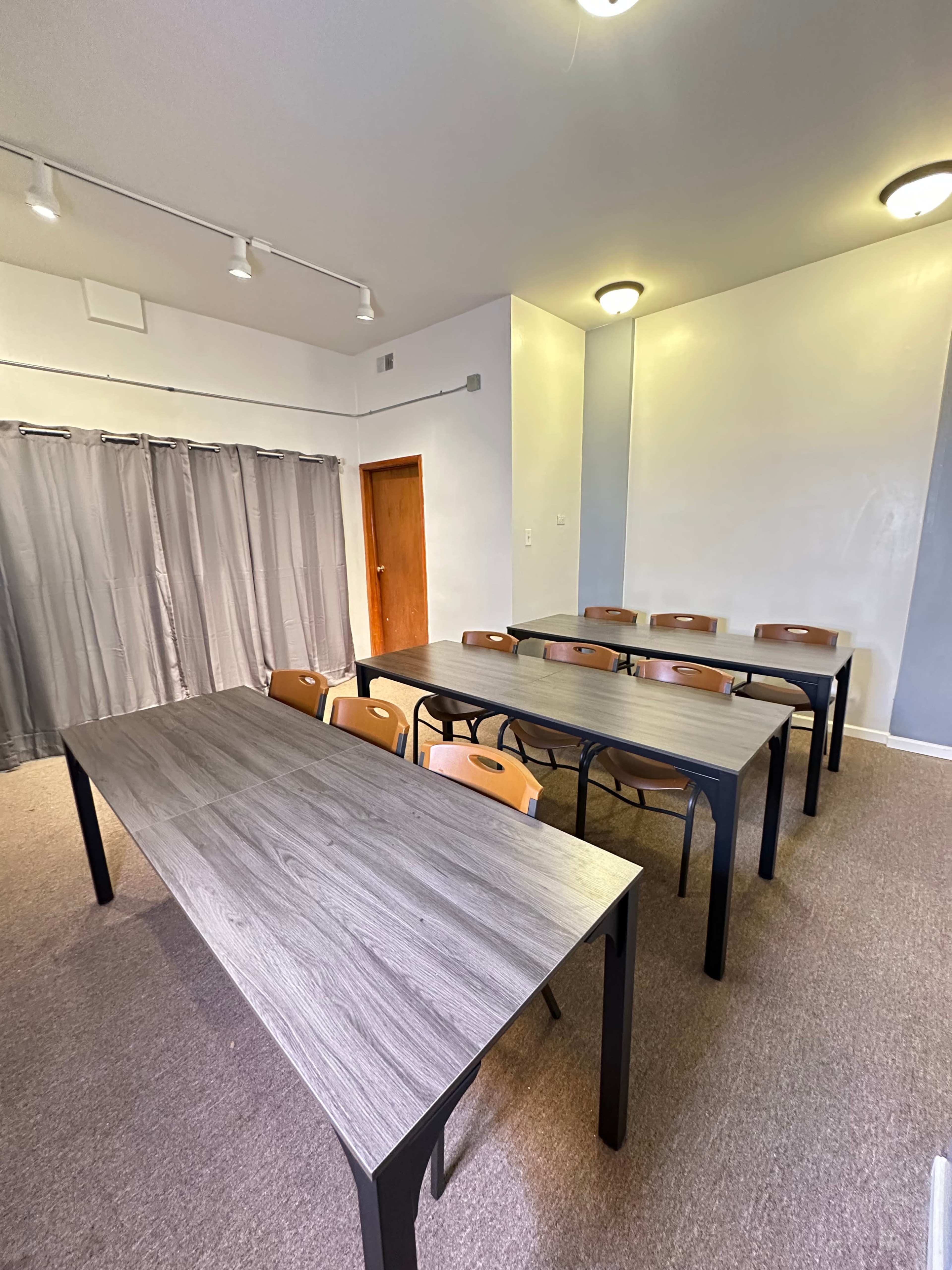 The image shows a classroom with two rows of dark wooden tables and chairs, along with gray curtains on one side and a wooden door in the background.