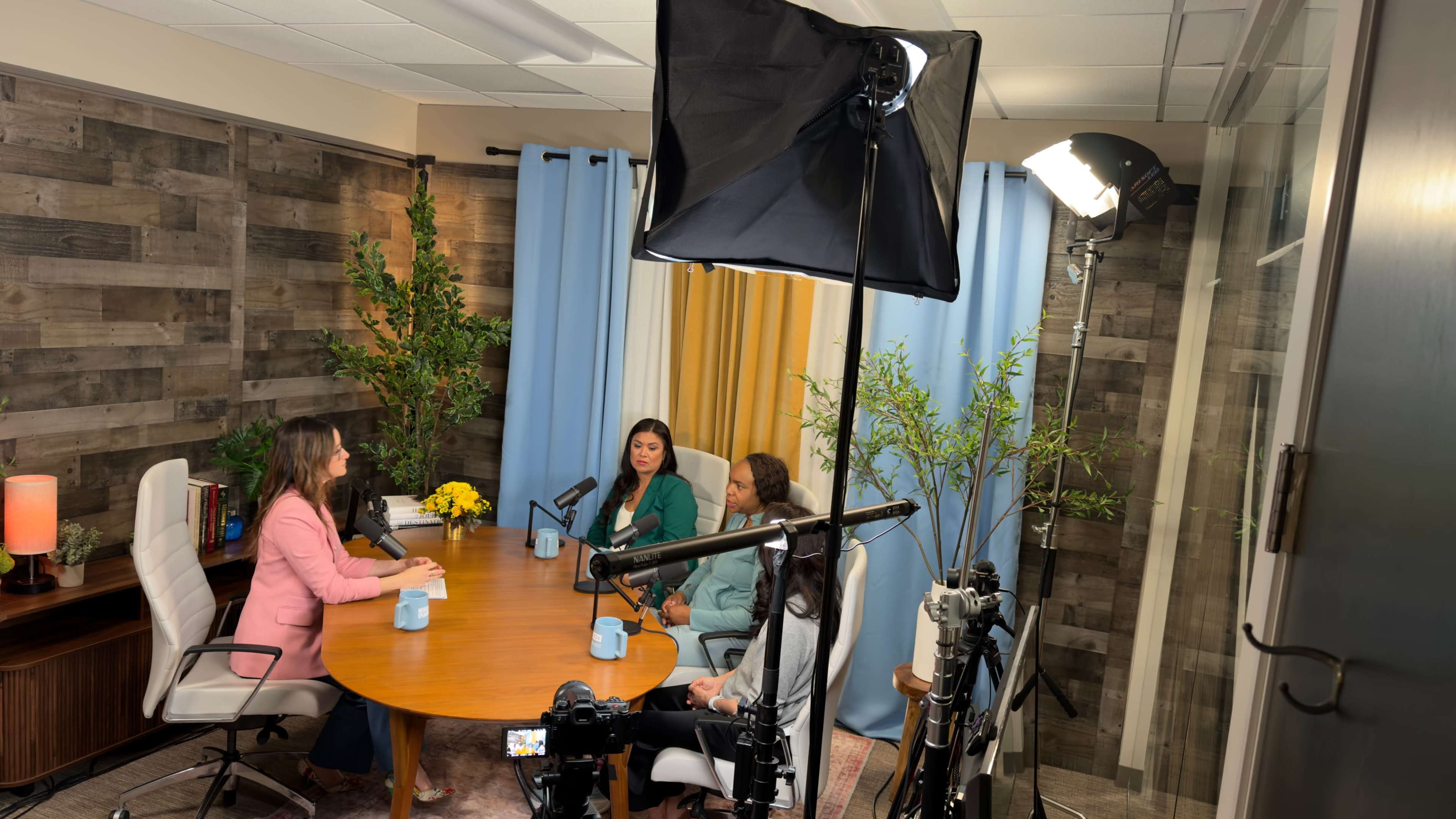 Three women are seated around a wooden table in a modern office space, while a camera and lighting equipment are set up to record a discussion.